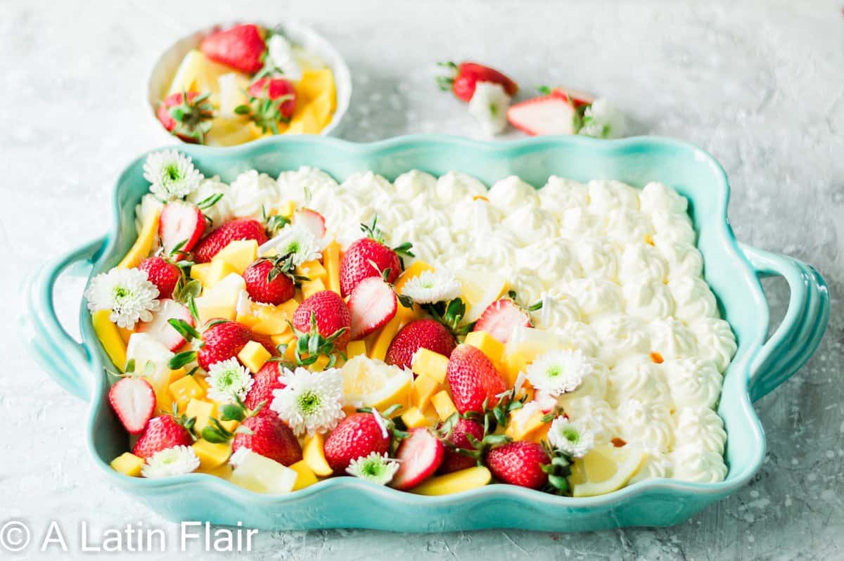 A vibrant dessert features a variety of fresh fruits such as strawberries and sliced mango, adorned with small white flowers and cream dollops, served in a large light blue baking dish, with additional strawberries and a small bowl of fruit in the background.