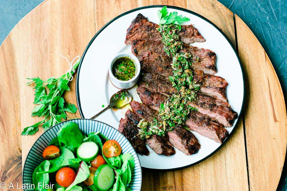 Skirt steak with chimichurri sauce drizzled and a side salad over a wood board