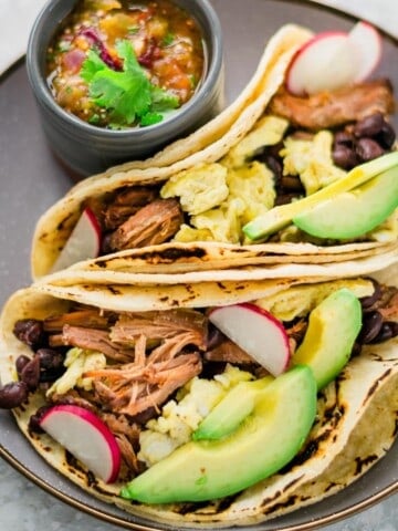 pork-carnitas-and-eggs-breakfast-tacos-with-black-beans,-avocados,-on-a-gray-plate-and-served-with-salsa-on-the-side