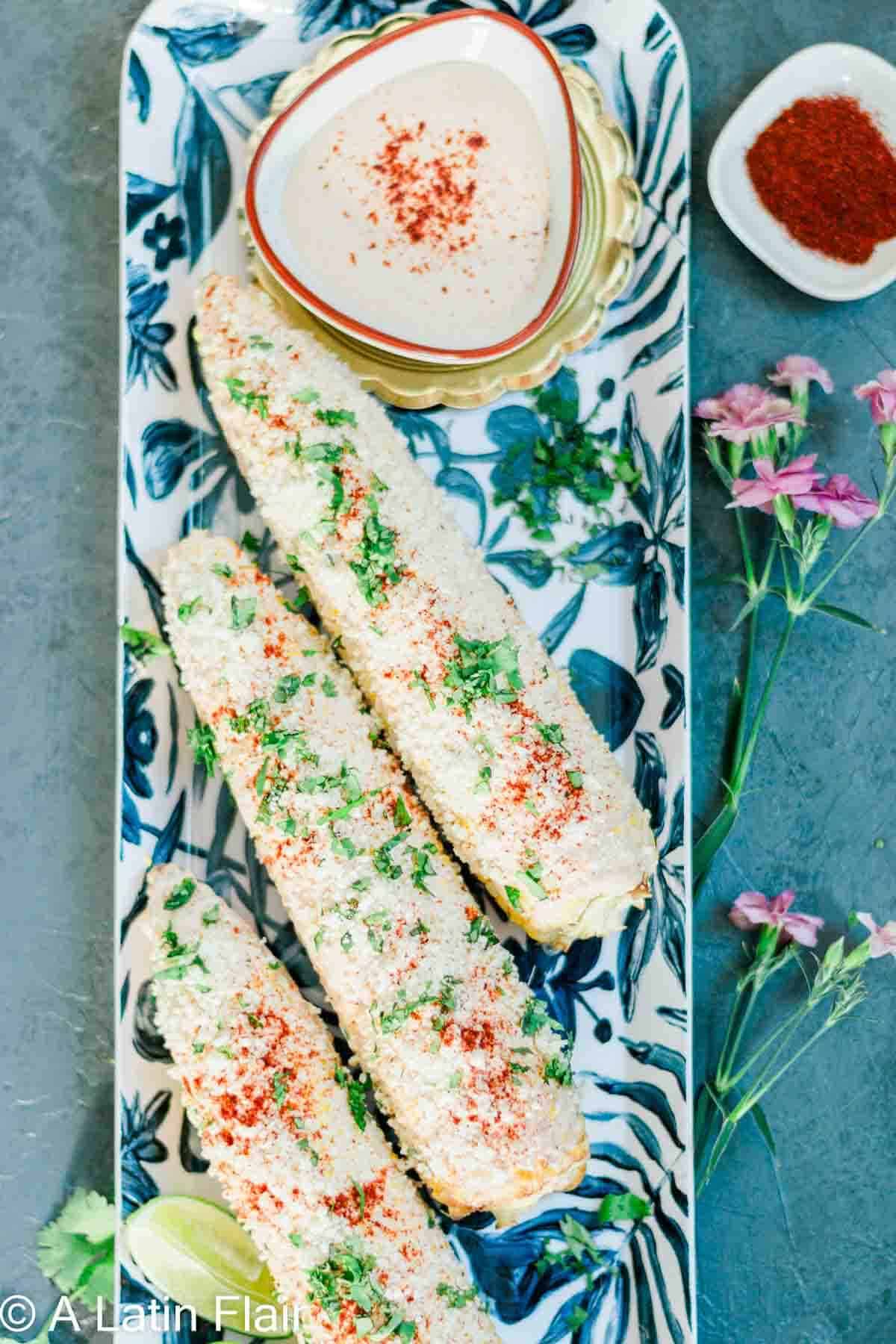 Three ears of Mexican street corn, or elote, covered in cotija cheese, cilantro, and chili powder on a floral platter. Lime wedge and pink flowers nearby.