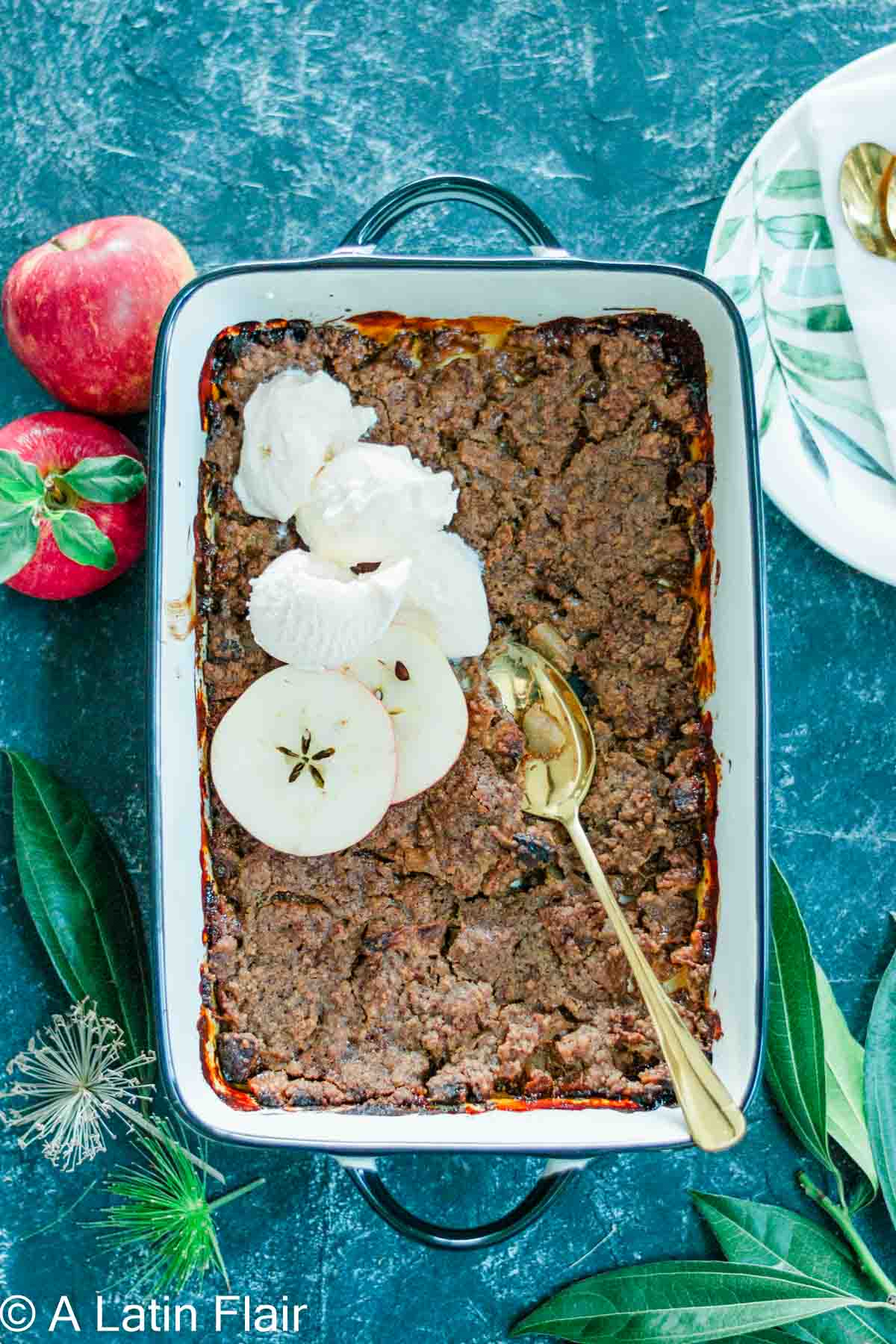 A baked apple crisp in a rectangular dish, topped with sliced apples and scoops of ice cream. A gold spoon is placed on top. Nearby are fresh red apples and green leaves, creating a cozy, autumnal feel.