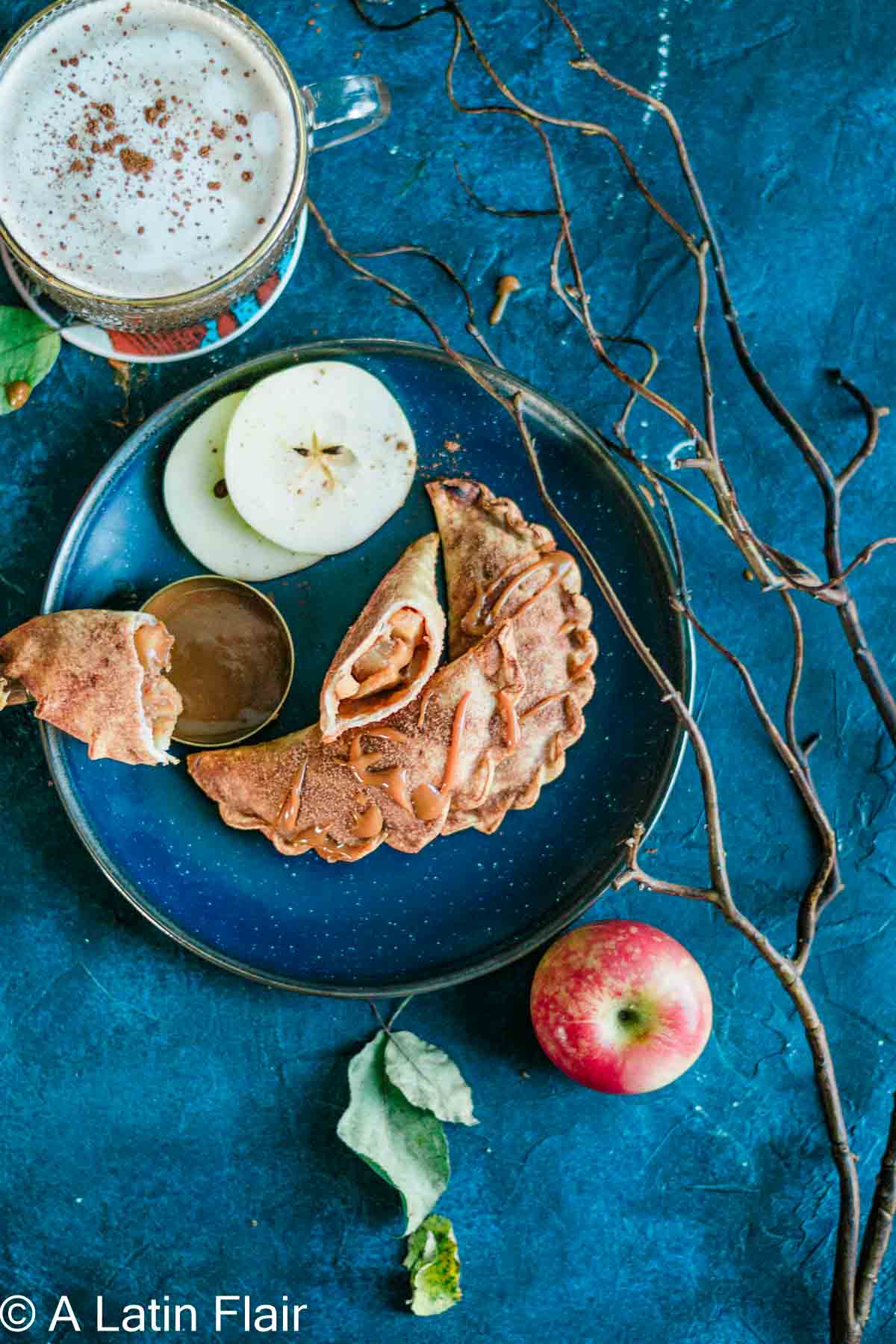 A blue plate with folded empanadas and apple slices, accompanied by a small cup of caramel, sits beside a frothy drink and an apple on a textured blue surface. Twigs surround the scene, adding a rustic feel.