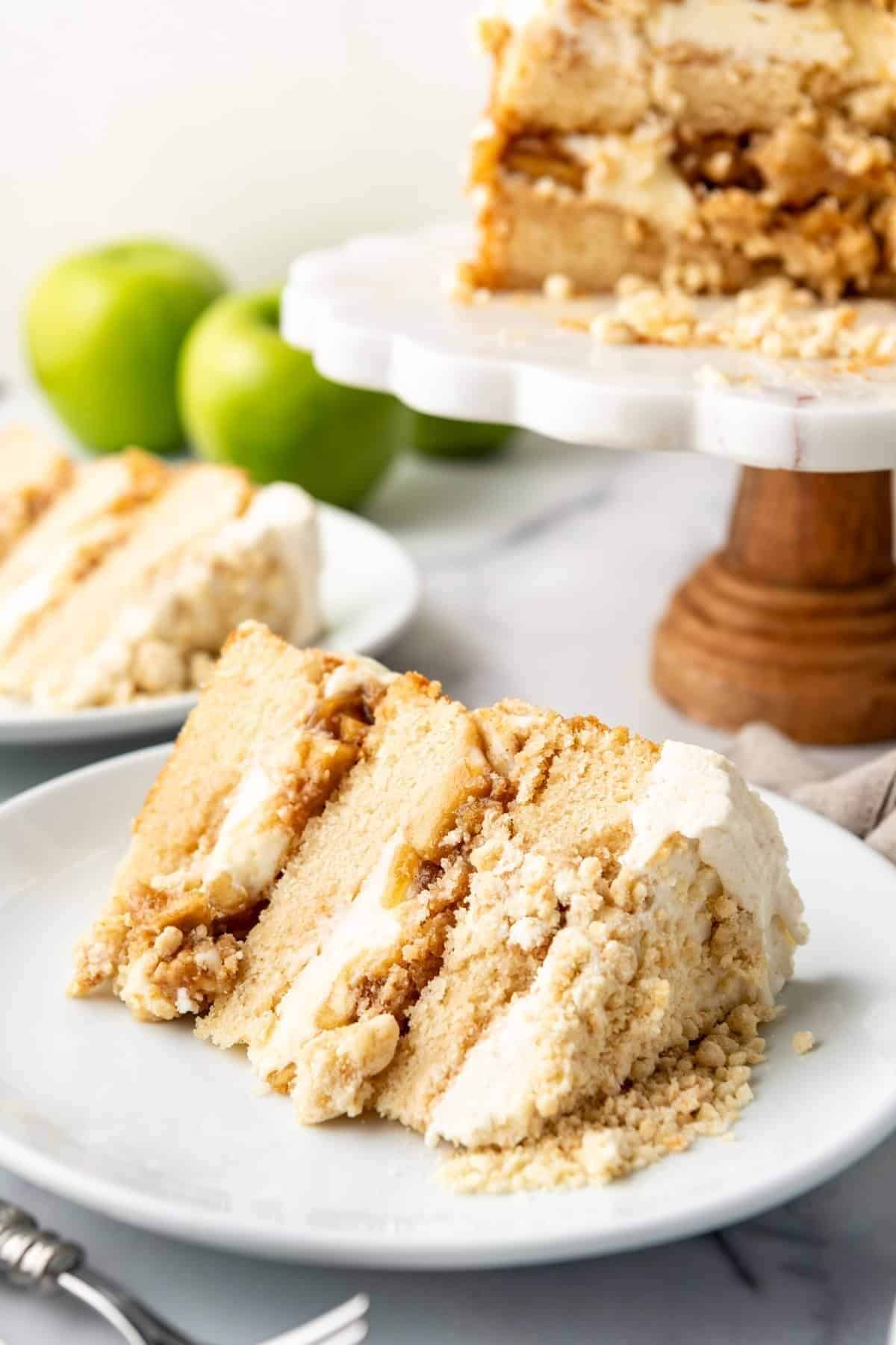 A slice of apple cake with creamy filling and crumb topping is on a white plate. A cake stand holds the remaining cake, with green apples nearby.