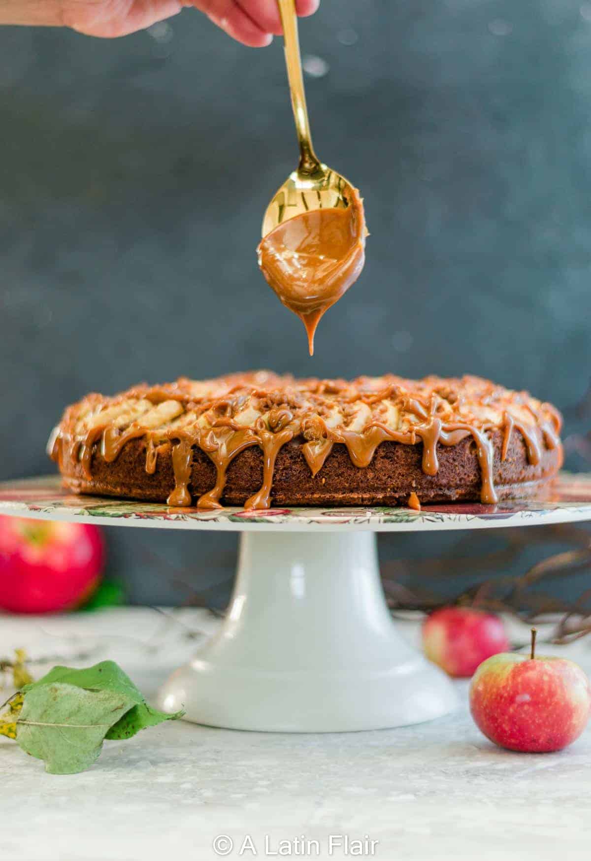 A rich caramel drizzle cascades over a dessert on a white stand, with apples in the blurred background, creating a warm and inviting atmosphere.