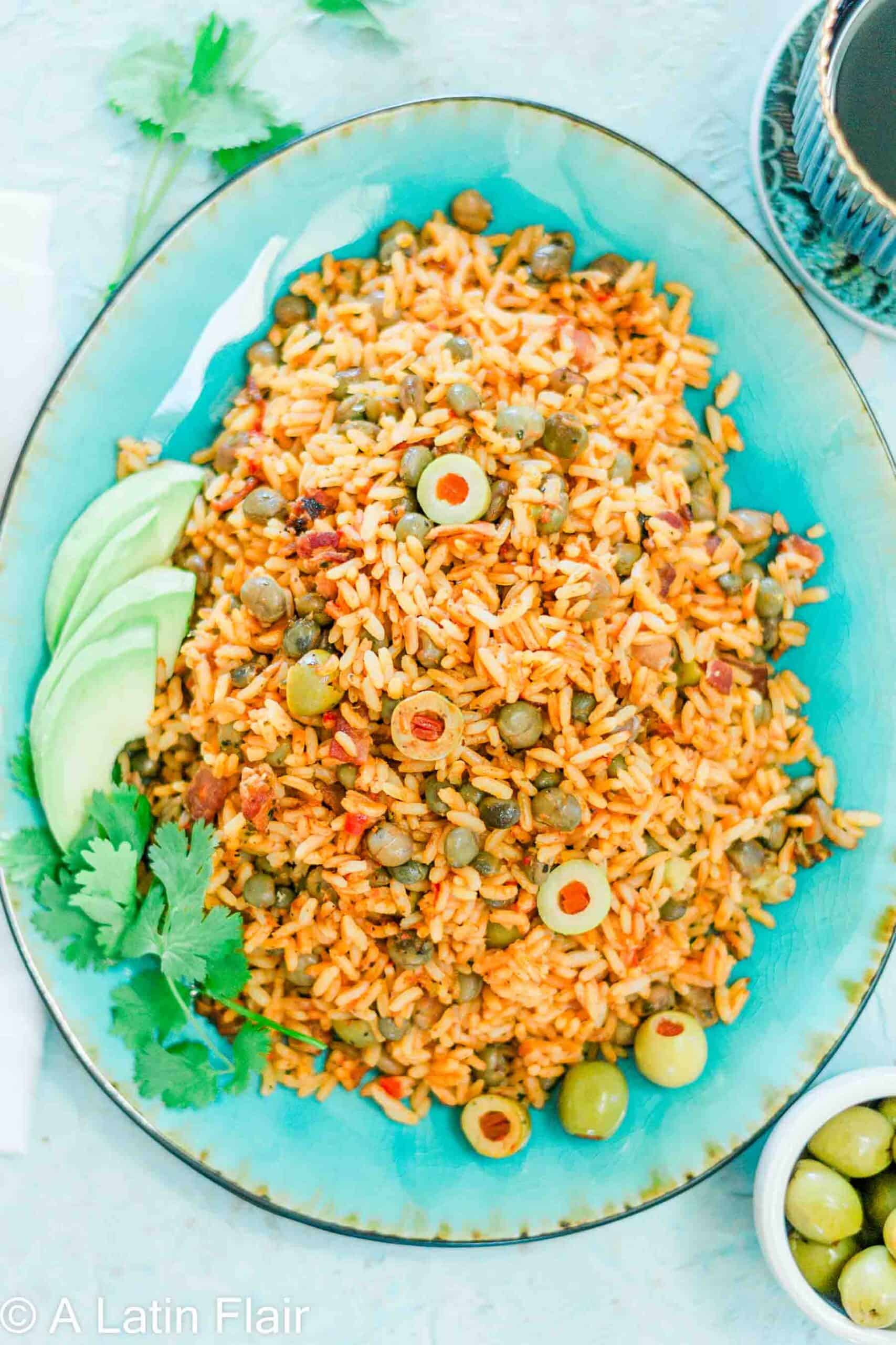 A vibrant turquoise plate filled with flavorful rice mixed with green olives, peas, and spices. Garnished with fresh avocado slices and cilantro.