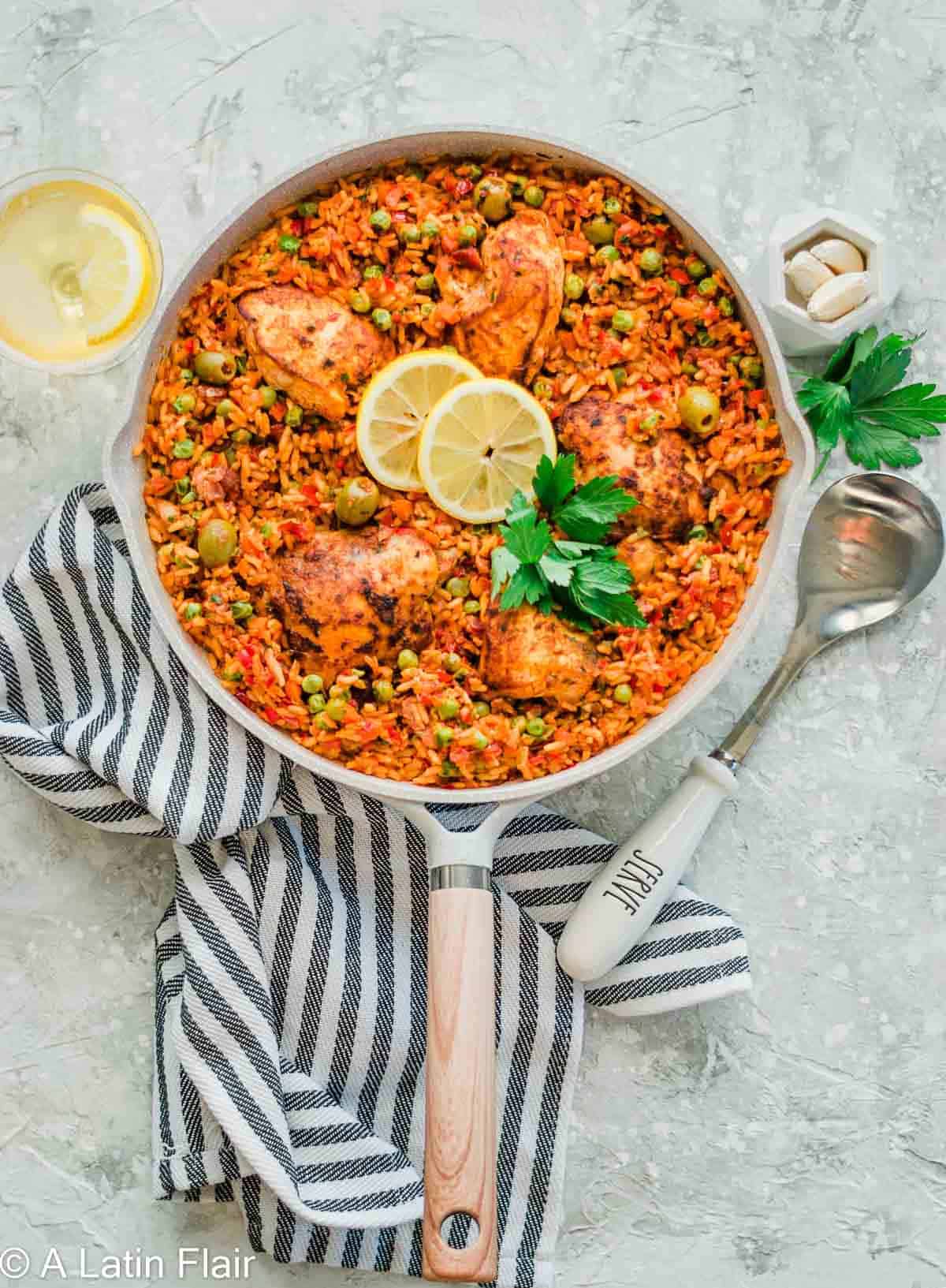 A skillet of vibrant arroz con pollo with golden chicken, peas, and olives, garnished with lemon slices and parsley. A striped towel and lemon slice beside.