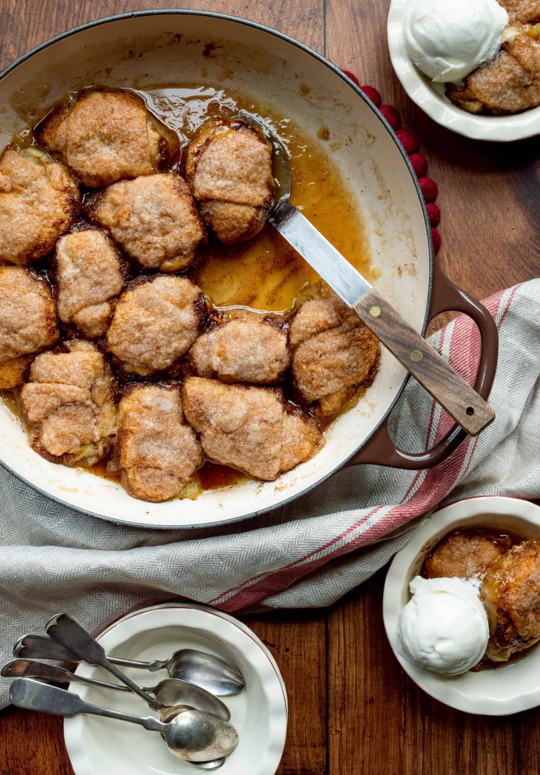 A warm cobbler with golden-brown biscuit topping fills a round baking dish, surrounded by a red-striped towel. Served with ice cream in bowls.