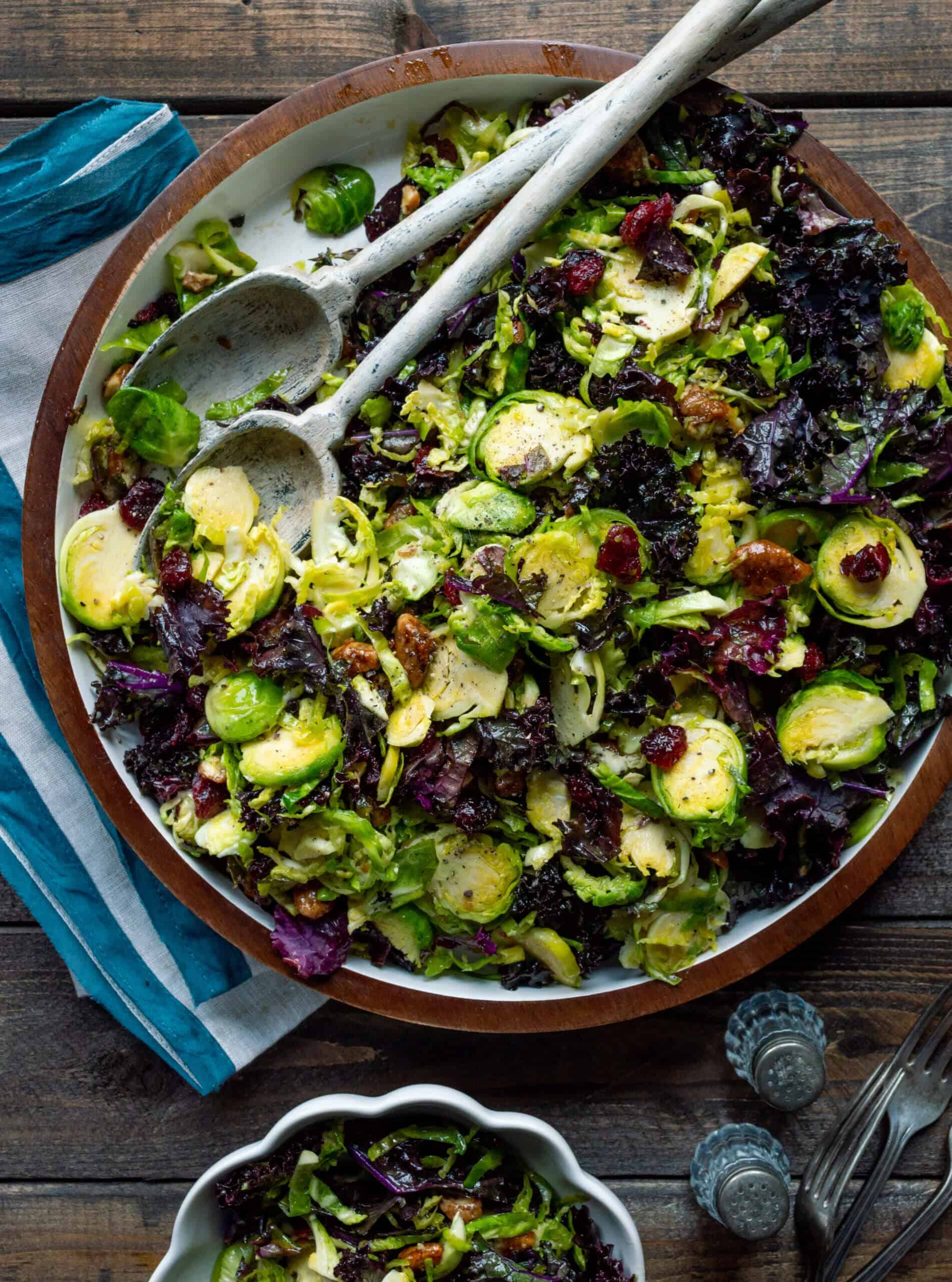A vibrant salad with Brussels sprouts, dark leafy greens, cranberries, and nuts in a wooden bowl. Wooden utensils and a striped cloth are beside it.