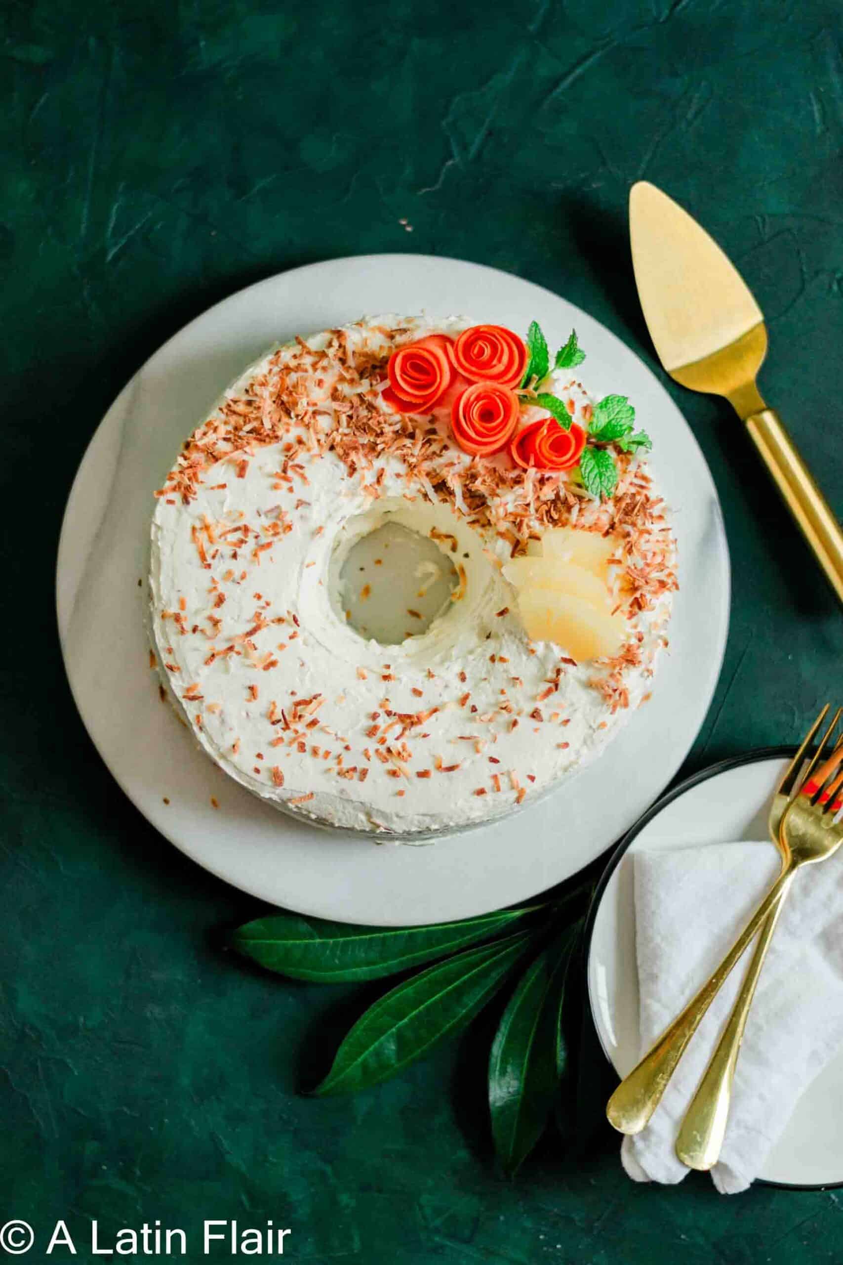 A round cake on a white plate with white frosting and coconut flakes, topped with red rose decorations, mint leaves, and pineapple slices.