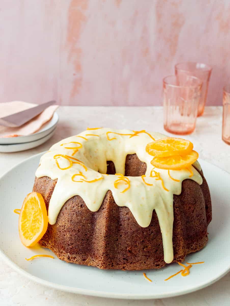 Bundt cake topped with white icing and orange zest, garnished with orange slices on a white plate. Soft pink background with stacked plates and glasses.