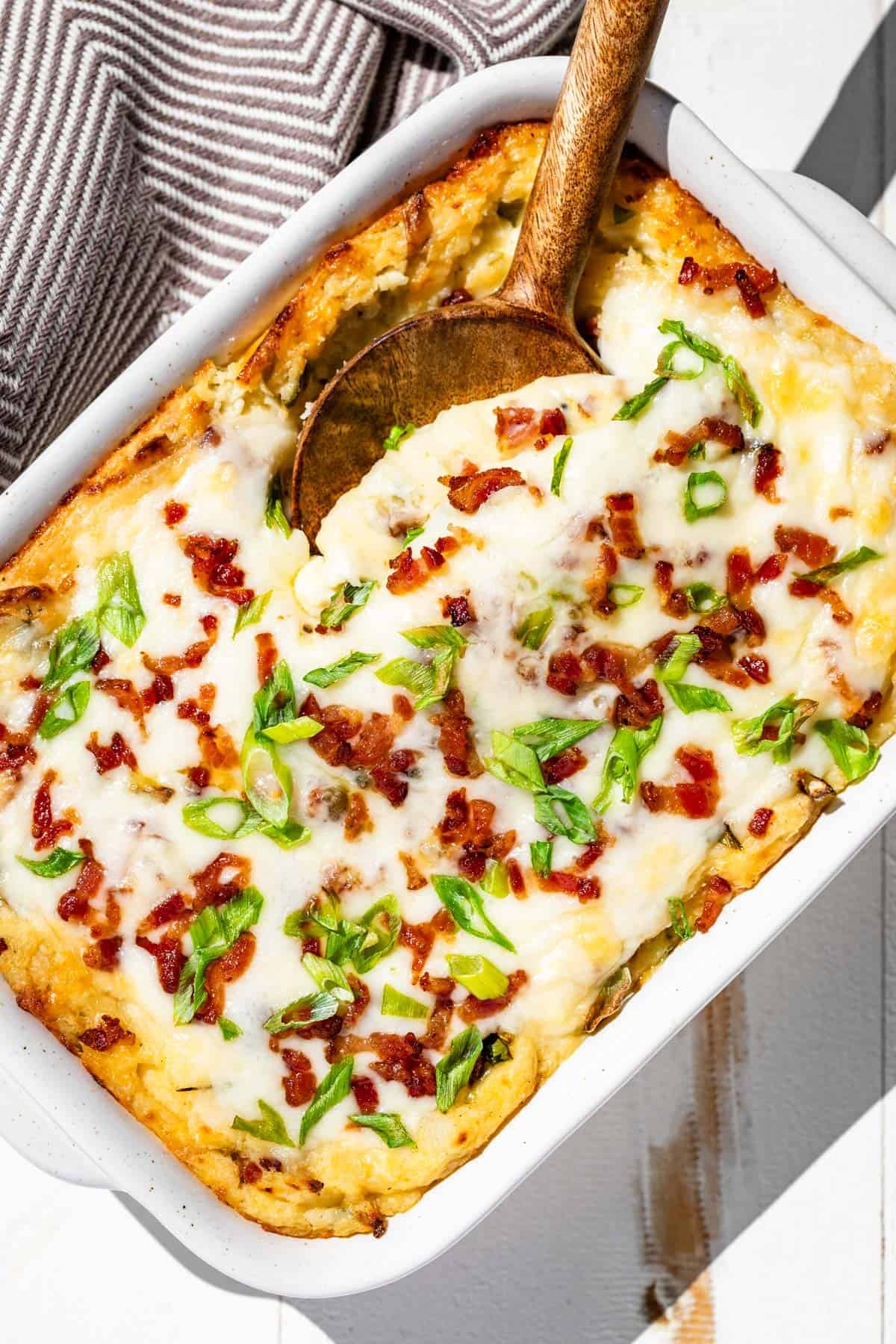 A baked casserole topped with melted cheese, crispy bacon bits, and chopped green onions. A wooden spoon rests in the dish, suggesting it's ready to serve.