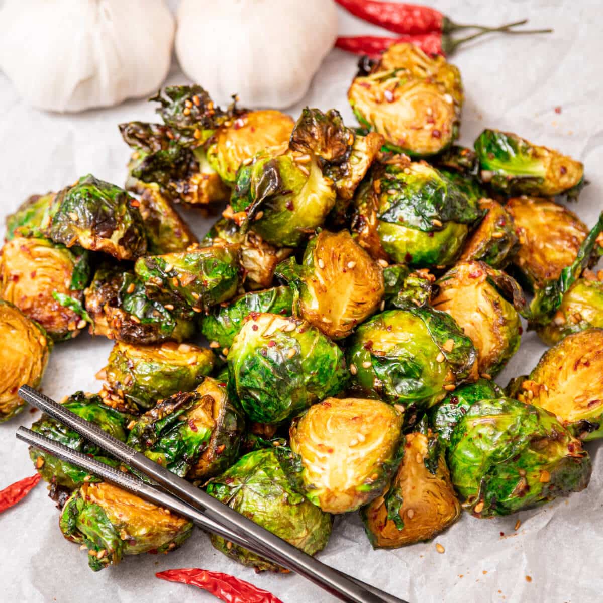 Roasted Brussels sprouts with a spicy glaze, garnished with sesame seeds and chili flakes. Two garlic bulbs and red chili peppers are in the background.