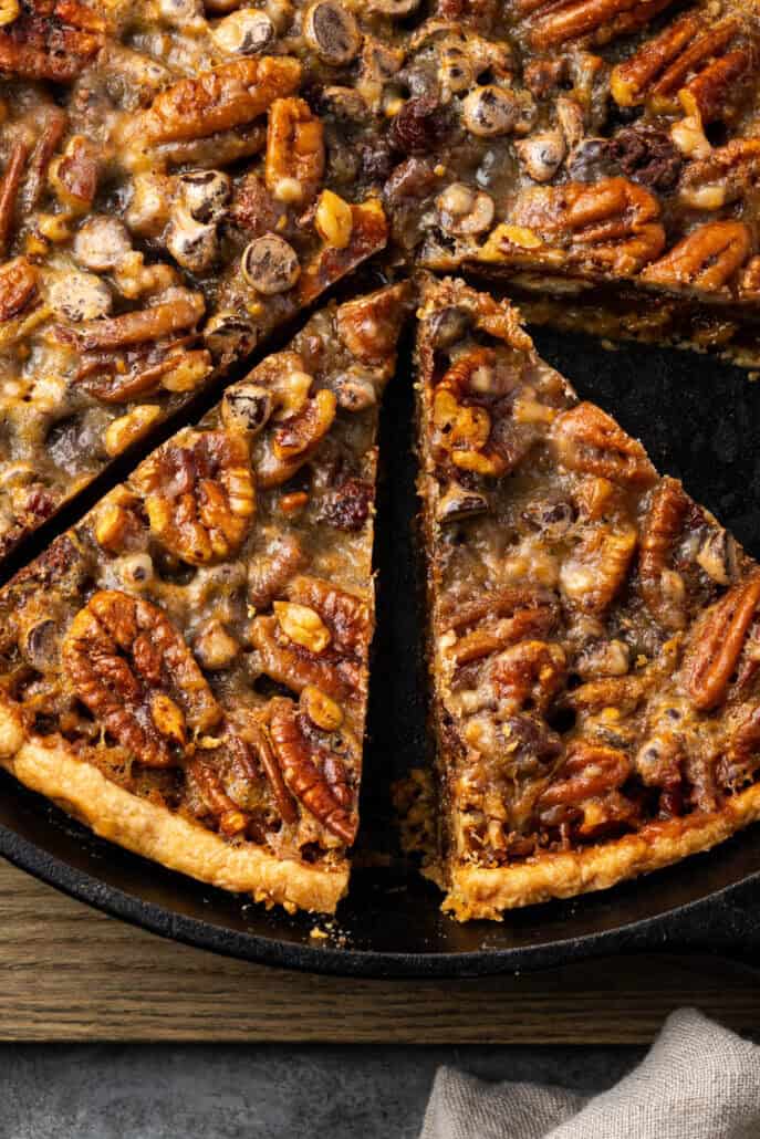 Close-up of a pecan pie with a slice taken out. The pie is richly topped with pecans and chocolate chips, sitting on a dark pan and wooden surface.