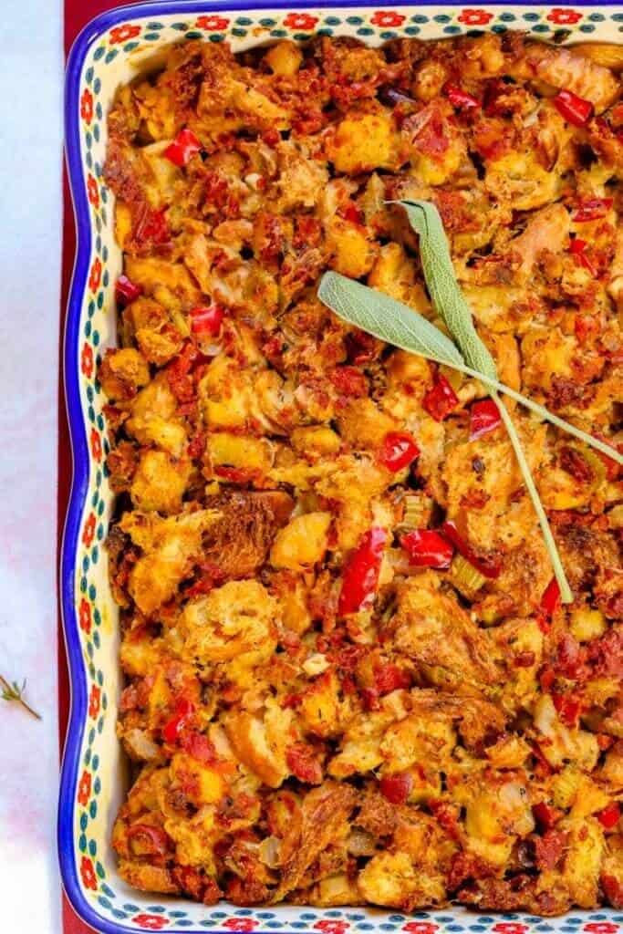 Casserole dish with baked stuffing, featuring golden-brown bread chunks, red bell peppers, and herbs. Topped with two sage leaves, giving a hearty, rustic look.