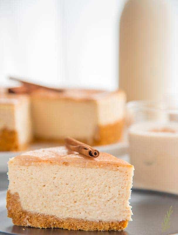 A slice of creamy cheesecake with a golden-brown crust is topped with a cinnamon stick. The background features more cheesecake.