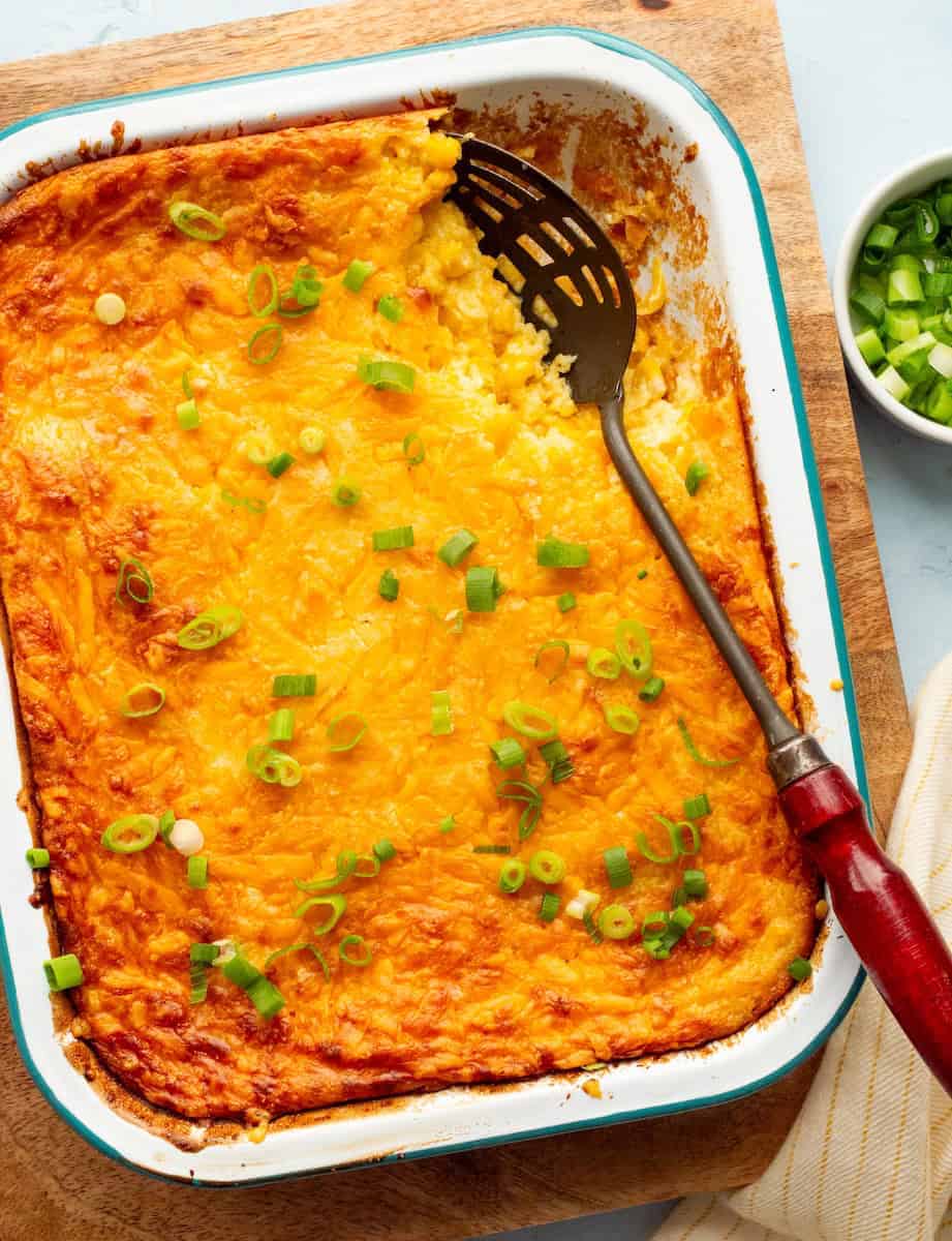 Golden, cheesy casserole topped with chopped green onions in a baking dish, partially served with a red-handled spatula. Cozy and appetizing look.