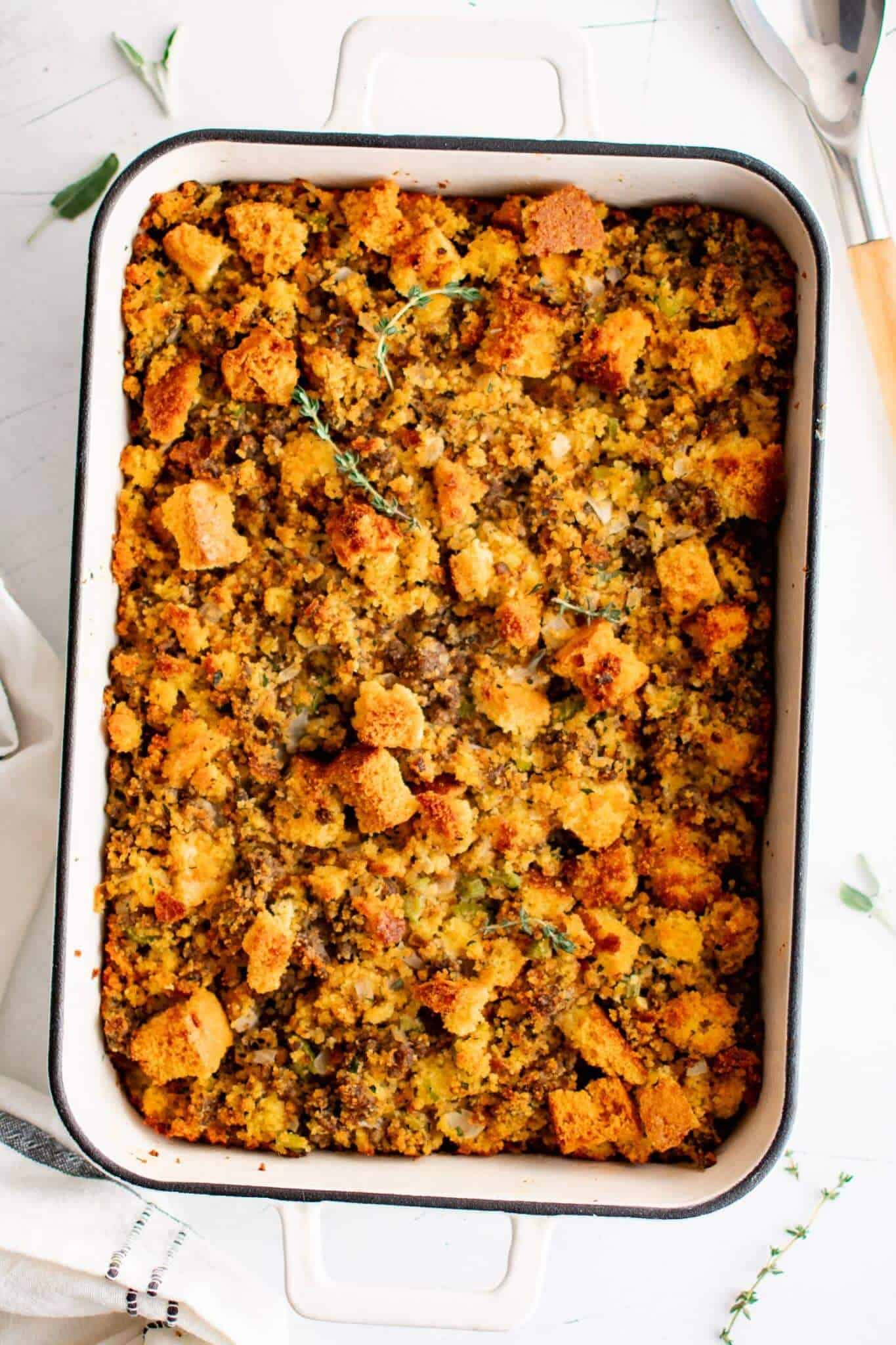 Golden-brown cornbread stuffing in a rectangular baking dish, topped with fresh herbs. The stuffing looks crispy and savory, conveying warmth.