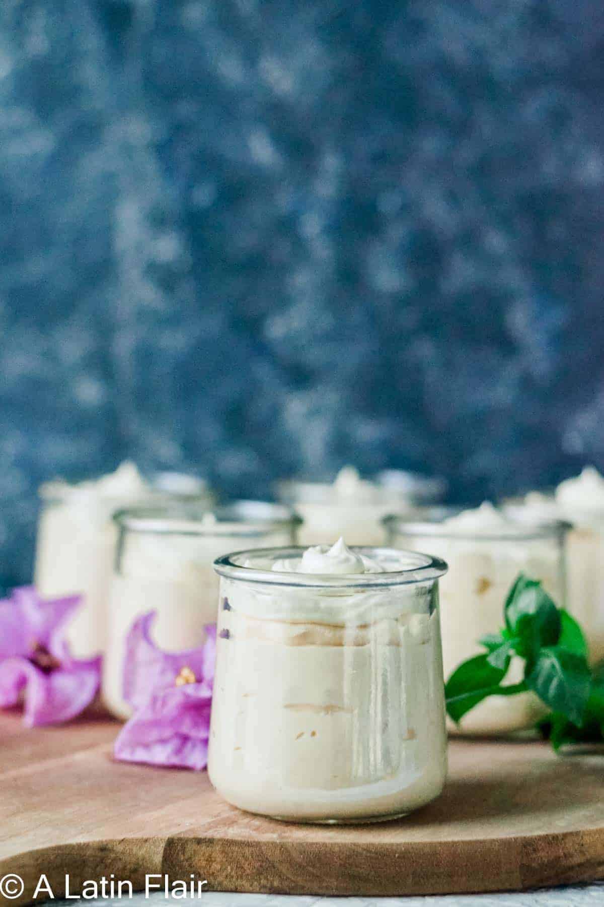 Creamy dessert in glass jars on a wooden board, surrounded by purple flowers and a sprig of green leaves, with a blurred blue background.