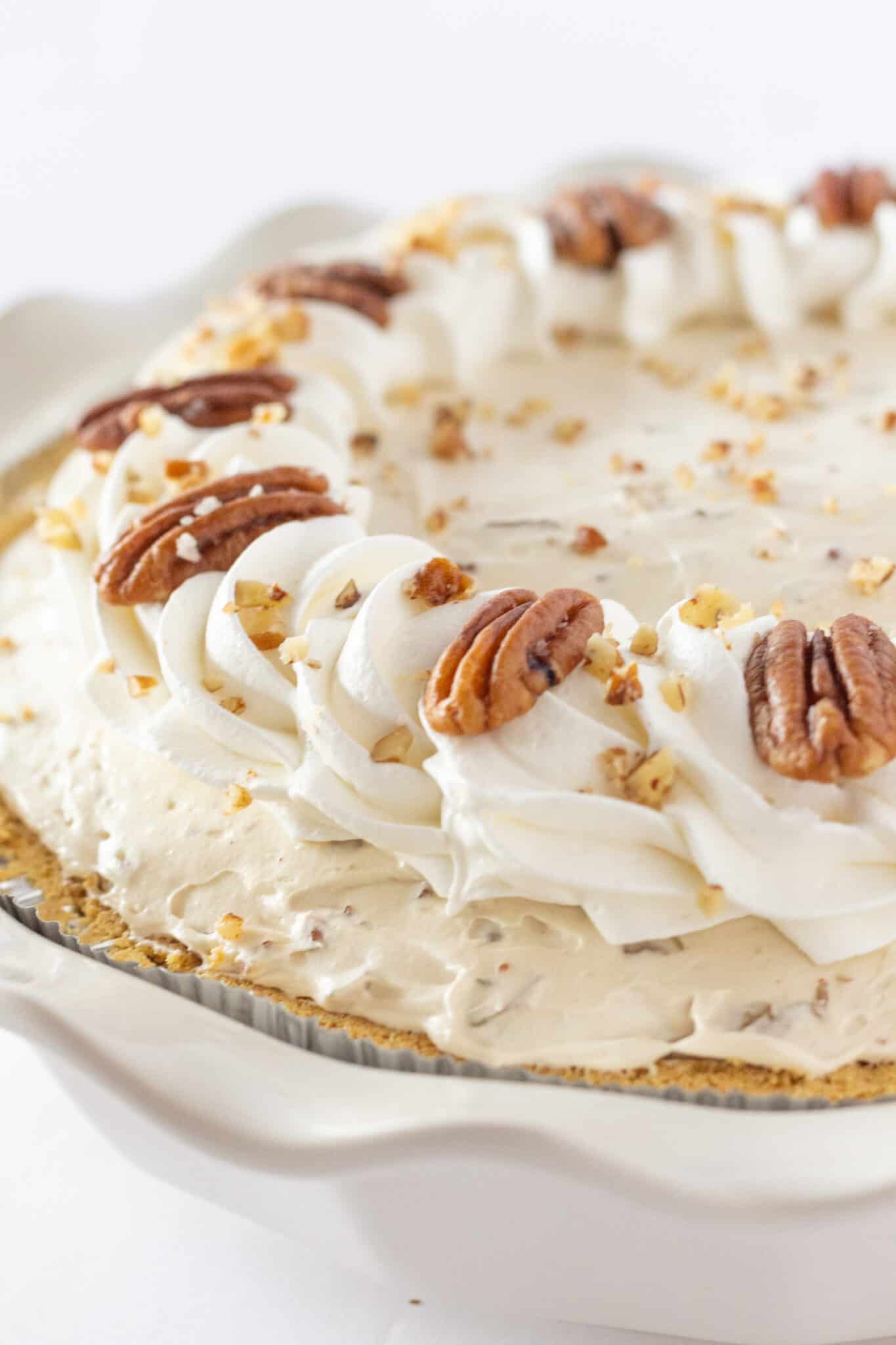 A creamy pie topped with whipped cream swirls, whole pecans, and crushed nuts. The pie crust is golden, creating a rich, inviting appearance.