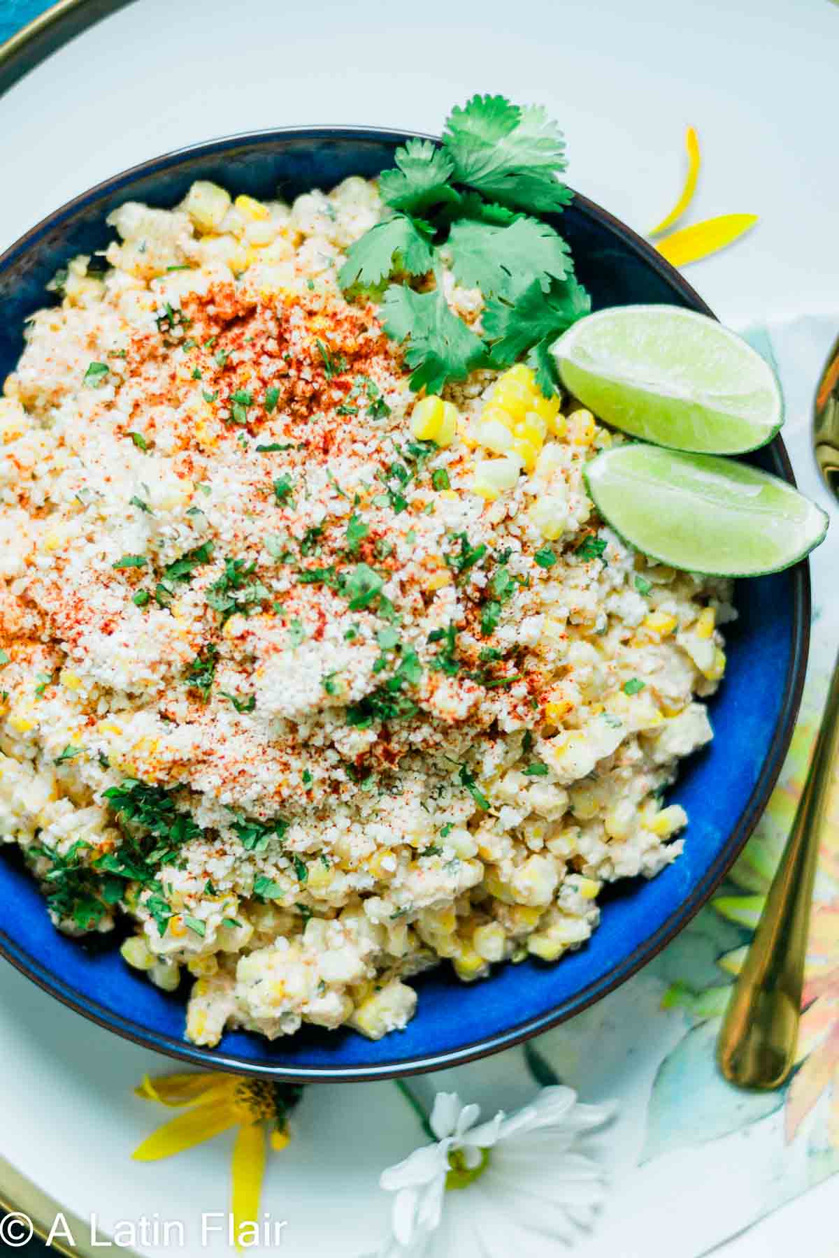 Close-up of Mexican street corn salad in a blue bowl, garnished with chili powder, cilantro, and lime wedges, on a floral-patterned surface.