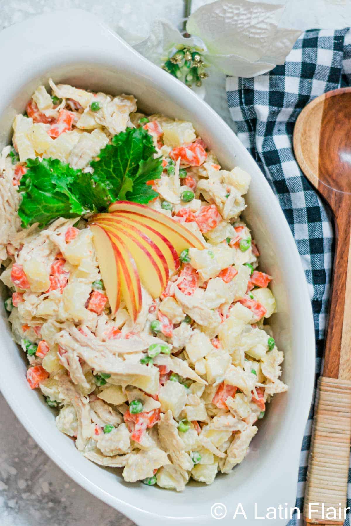 A creamy chicken potato salad served in a white oval dish, made with diced potatoes, shredded chicken, carrots, and green peas, garnished with fresh greens and thin apple slices — a colorful and hearty potato dish perfect for any occasion.