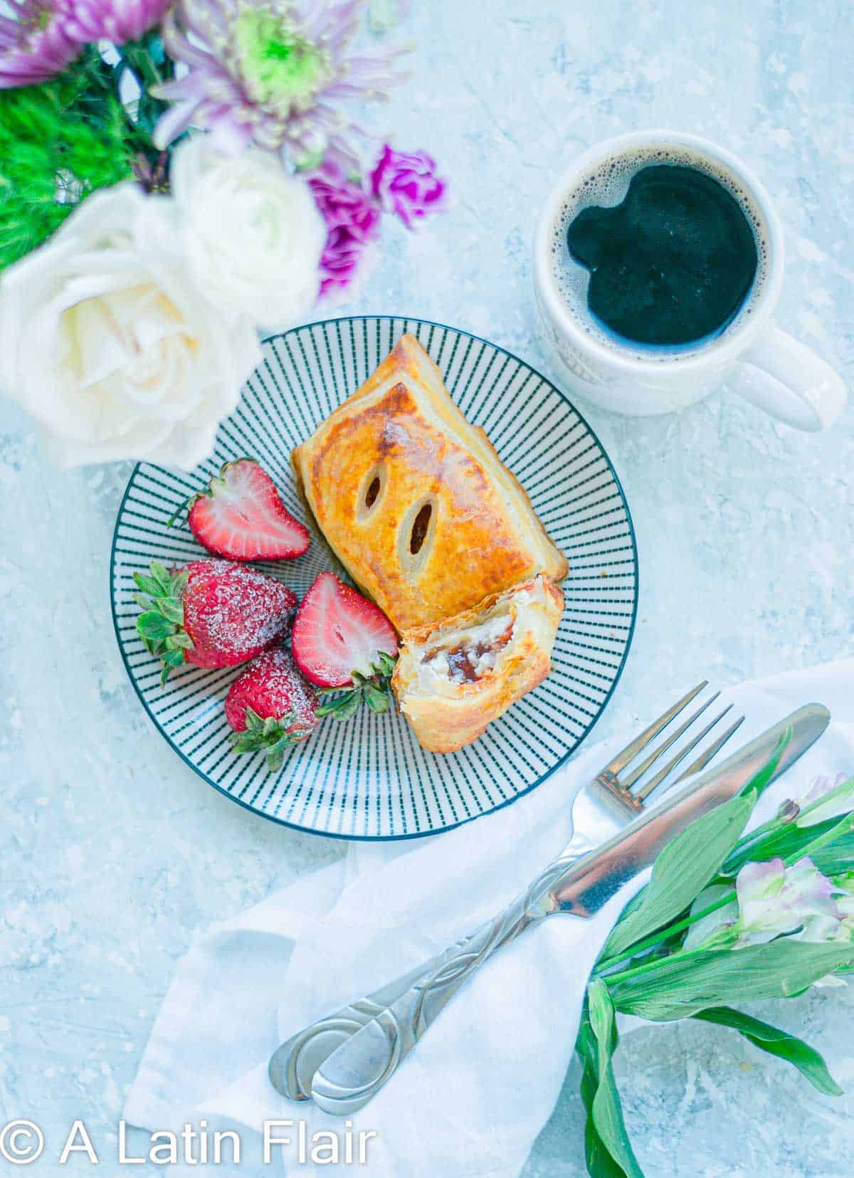 A plate with a flaky pastry and fresh strawberries, beside a white mug of black coffee. A bouquet of flowers and cutlery on a napkin are nearby.
