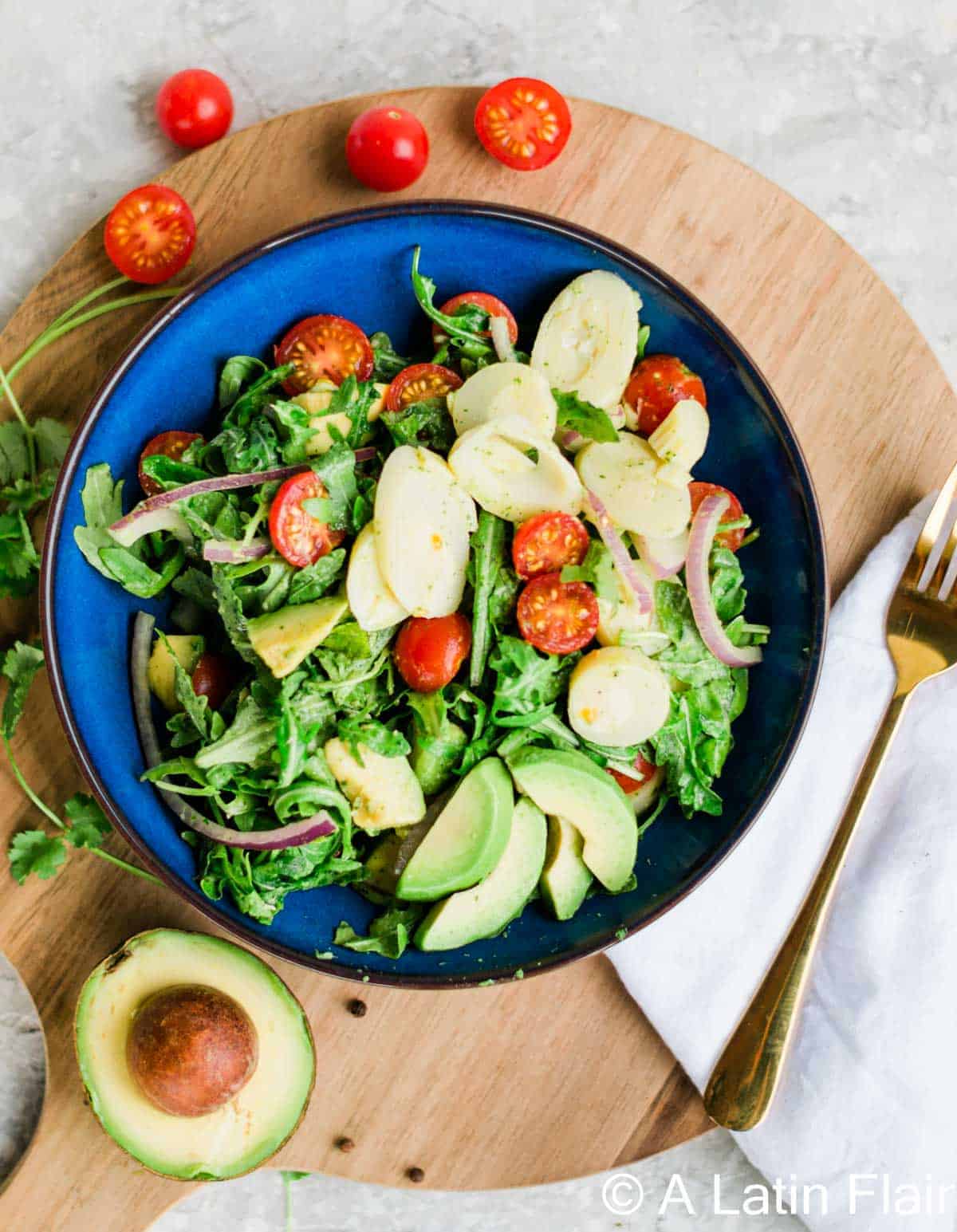 A vibrant salad with arugula, halved cherry tomatoes, avocado slices, hearts of palm, and red onion in a blue bowl on a wooden board. A fork and napkin are beside it.