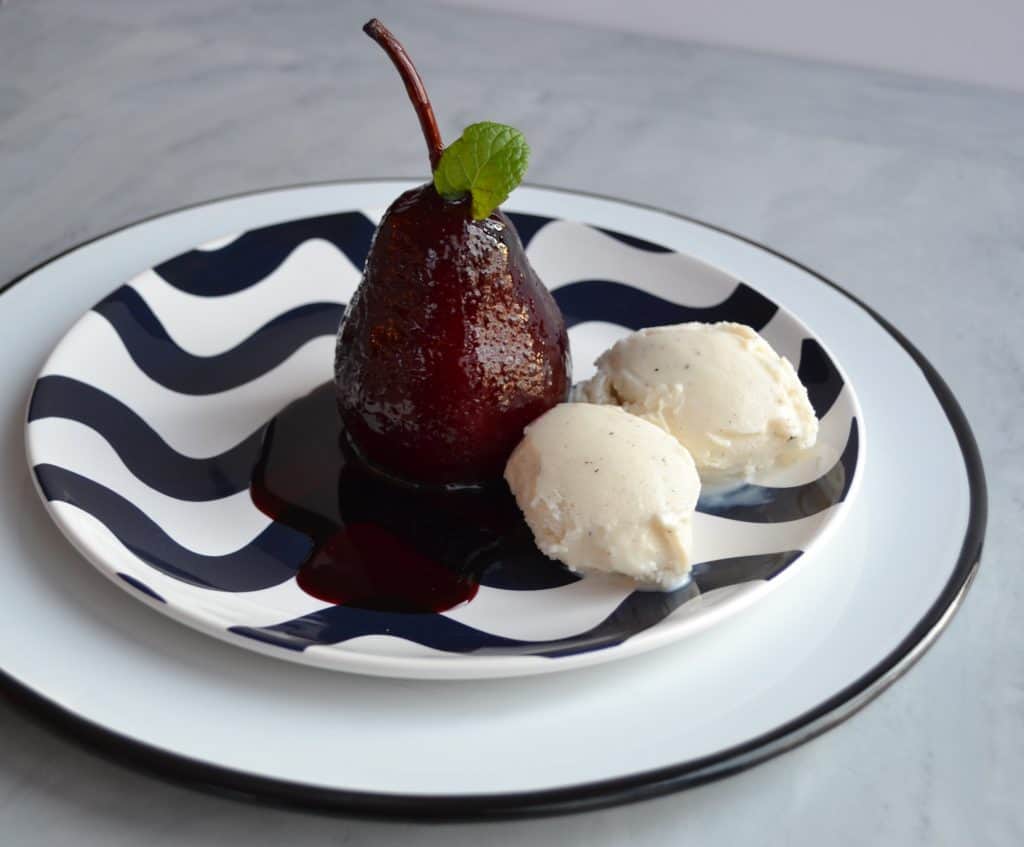A poached pear drizzled with dark sauce sits on a wavy-patterned plate alongside two scoops of vanilla ice cream, garnished with a mint leaf.