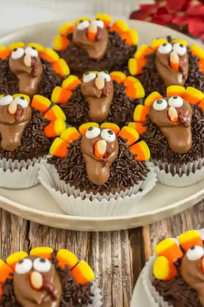 Cupcakes decorated as turkeys with chocolate frosting, candy corn feathers, and candy eyes, arranged on a plate on a rustic wooden table. Festive and playful tone.