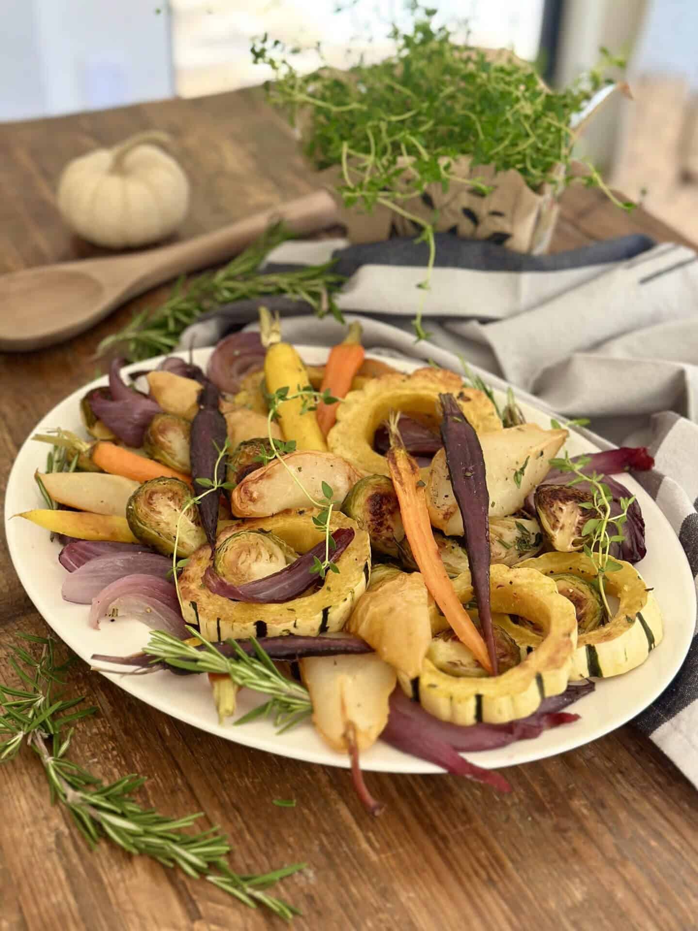 A platter of roasted vegetables including carrots, parsnips, brussels sprouts, and squash garnished with rosemary and thyme, on a wooden table.