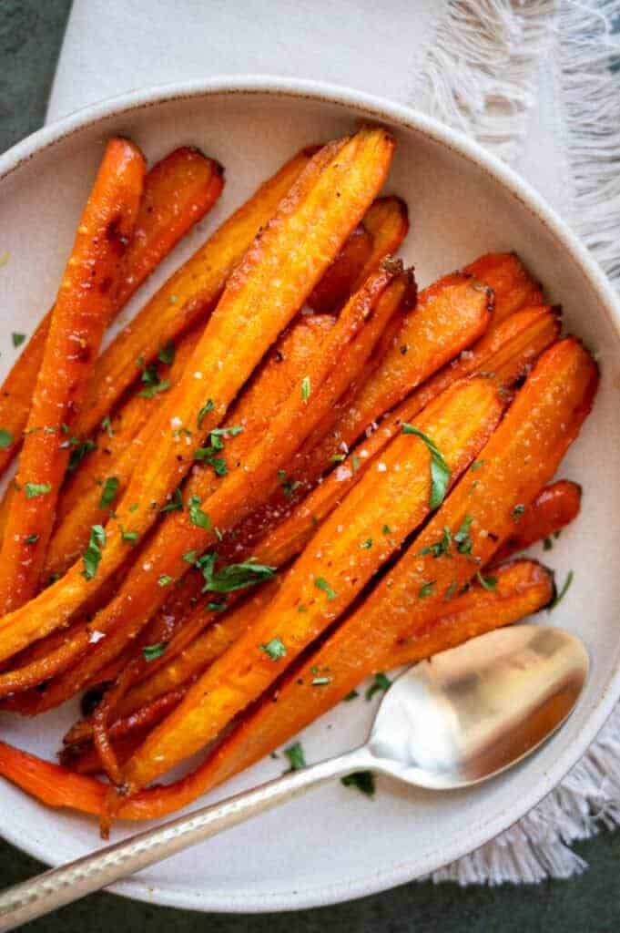 Roasted carrots on a white plate garnished with herbs, conveying a warm, appetizing tone. A silver spoon lies beside them on the fringed placemat.