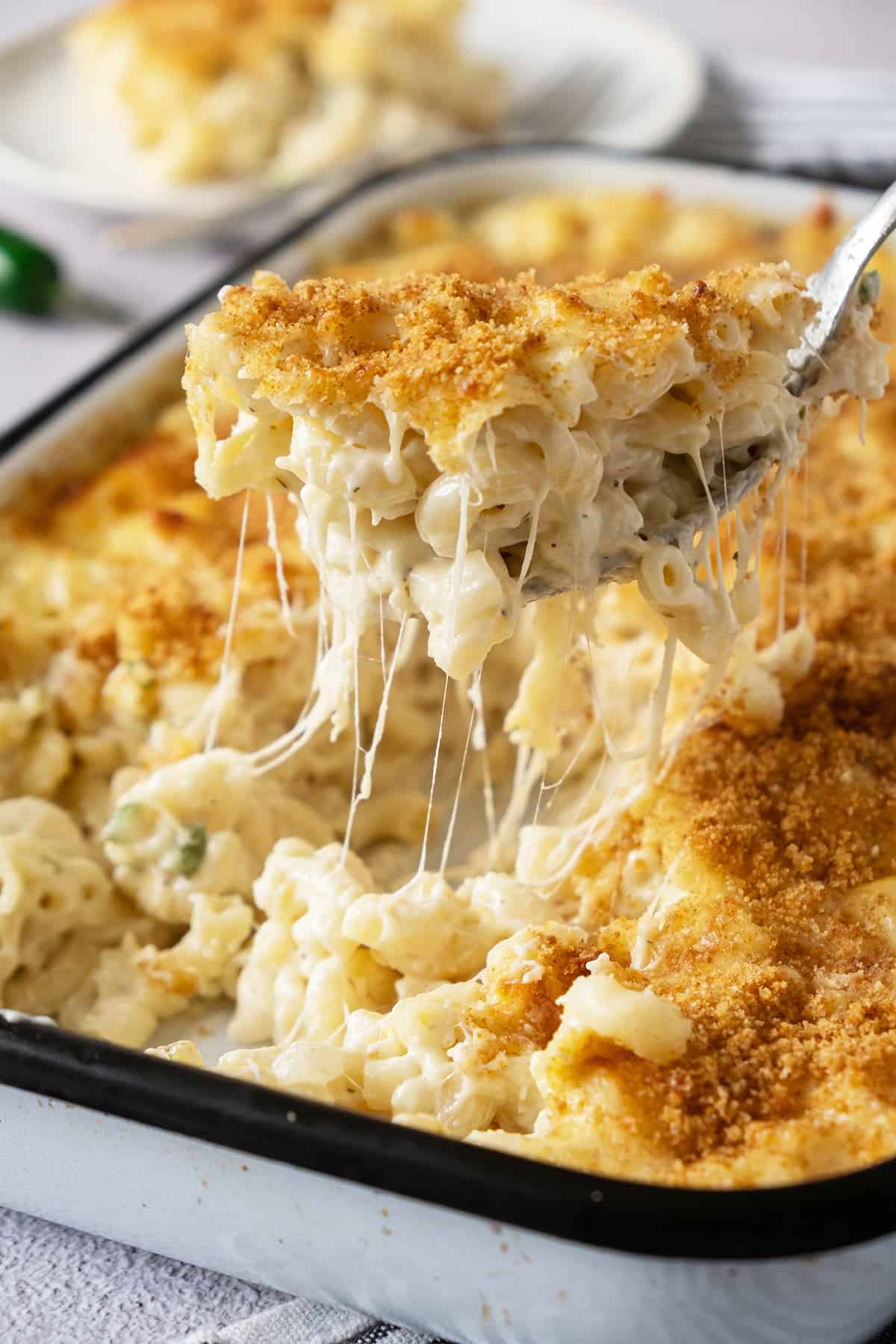 Close-up of a baked macaroni and cheese dish with a golden breadcrumb crust. A spoon lifts out a portion, showing gooey, melted cheese strands.