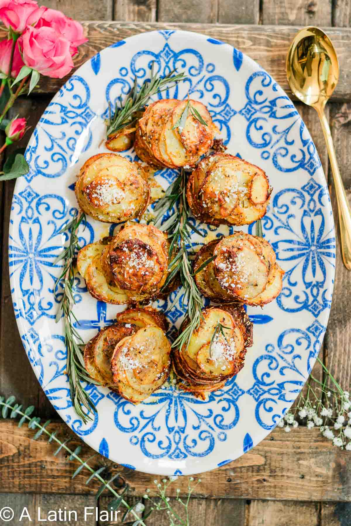 Ceramic platter with golden potato stacks garnished with rosemary, set on a rustic wooden table. A golden spoon and pink roses add elegance.