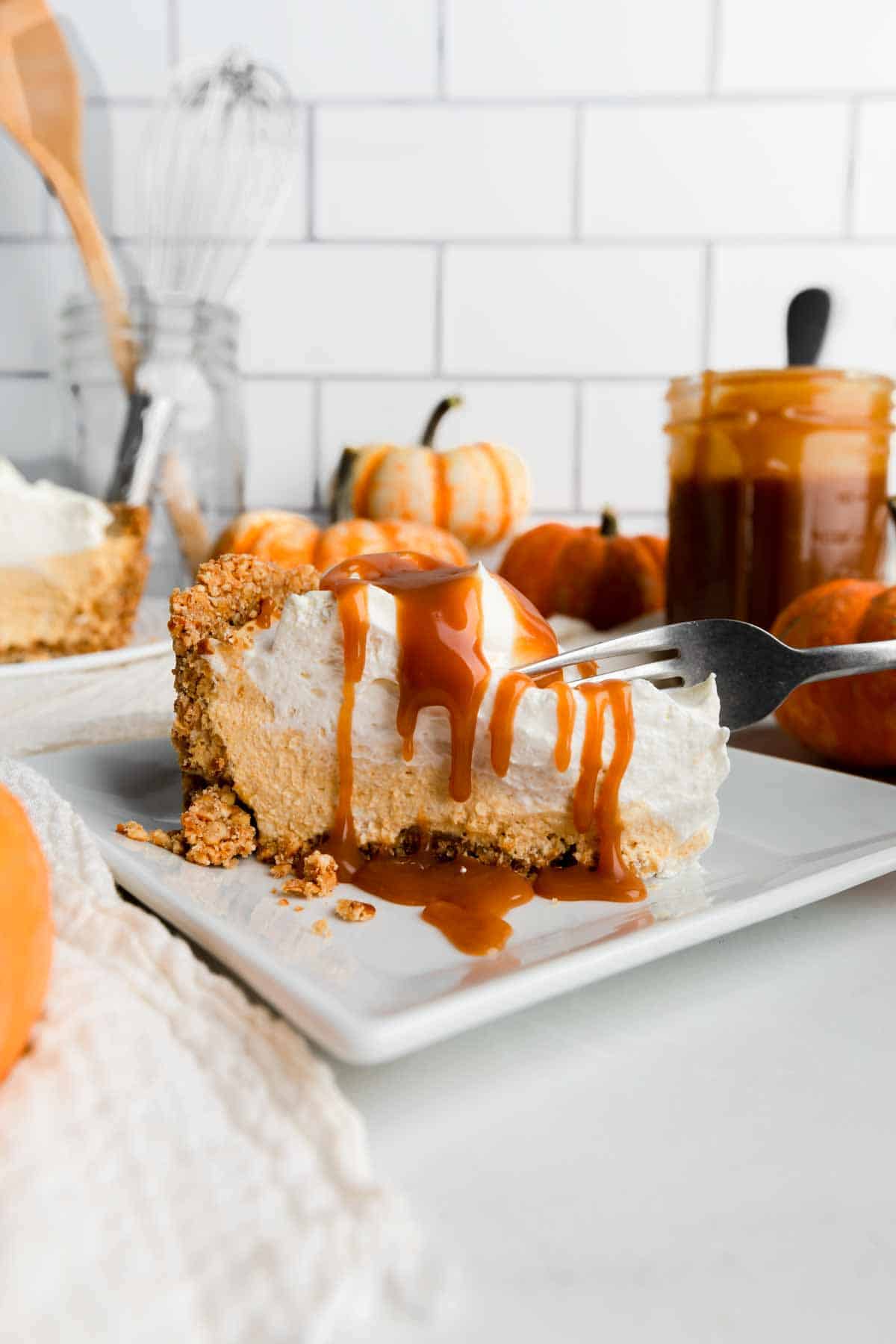A slice of pumpkin pie with whipped cream on a white plate, drizzled with caramel sauce. Mini pumpkins and a caramel jar in the background. Cozy and autumnal.