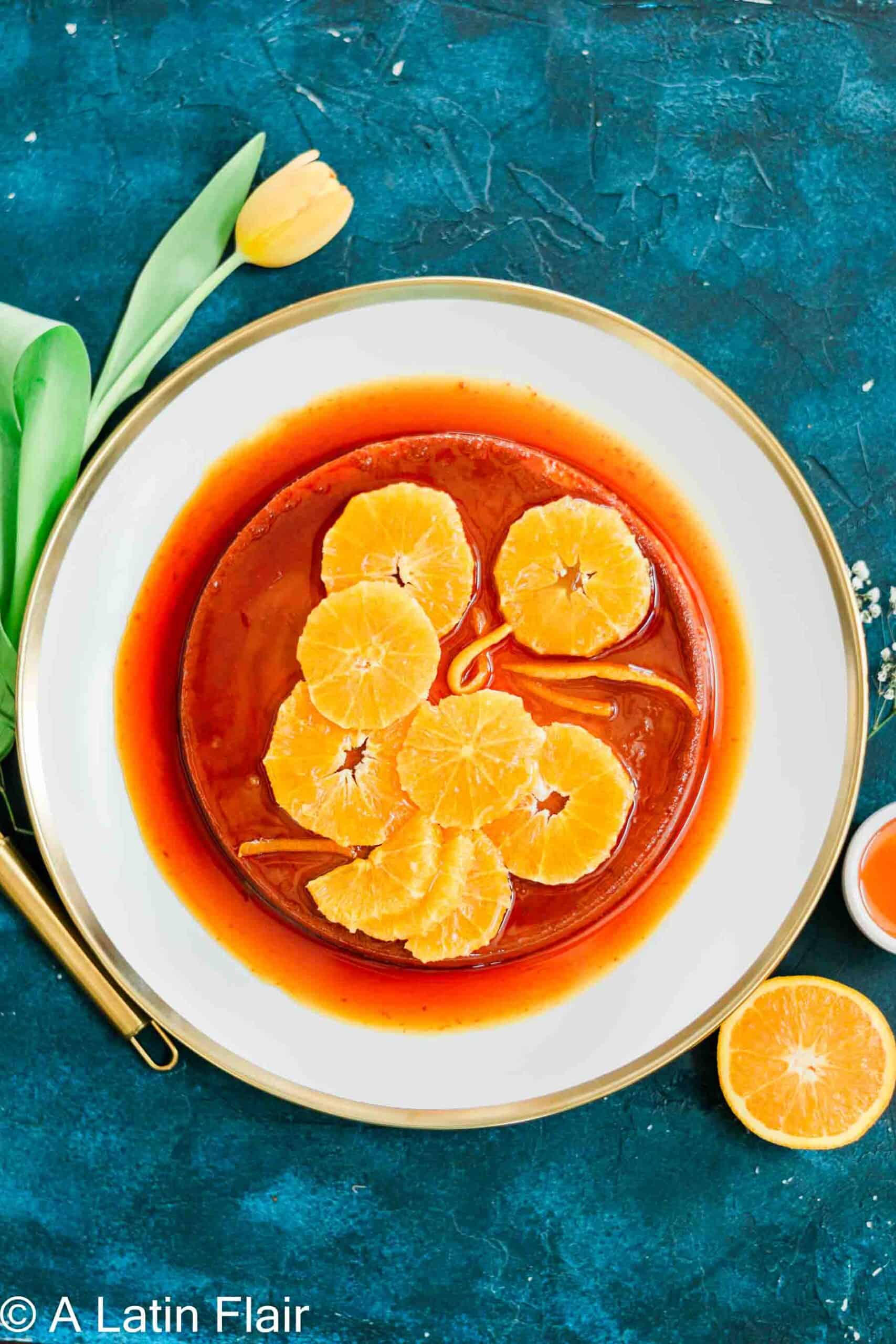 A round caramel flan topped with orange slices, sits on a white plate with a gold rim. A yellow tulip and orange halve are on a blue textured background.
