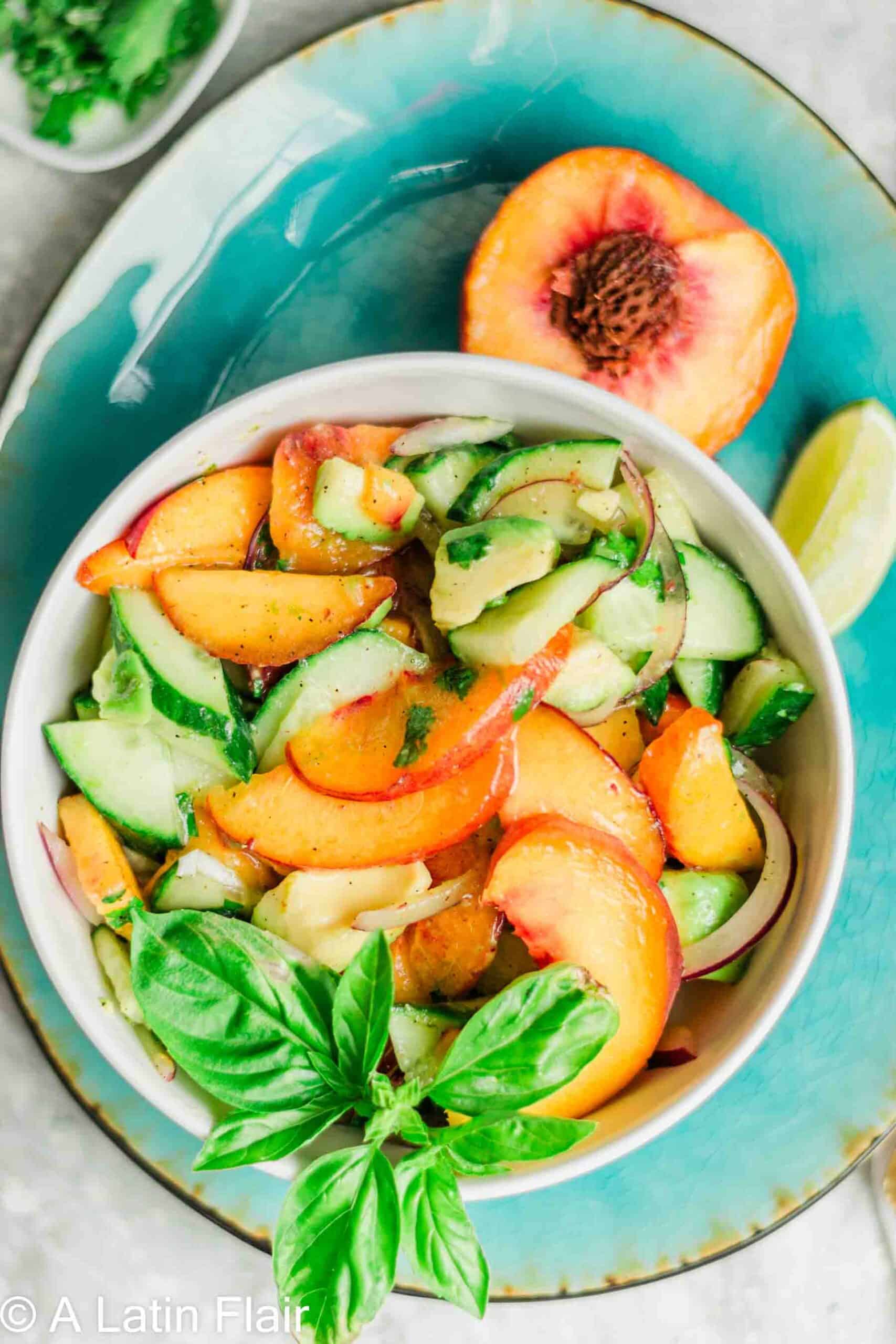 Fresh and vibrant salad with sliced peaches, cucumbers, and red onions in a white bowl garnished with basil. A halved peach and lime wedge on a teal plate add a summery feel.