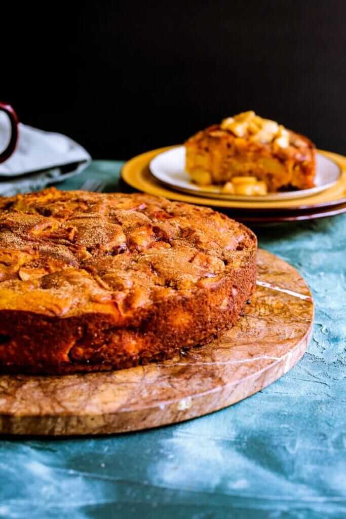 Golden-brown pumpkin pie on a marble board, with a slice on a yellow plate in the background. The tablecloth is teal, adding a warm, inviting feel.