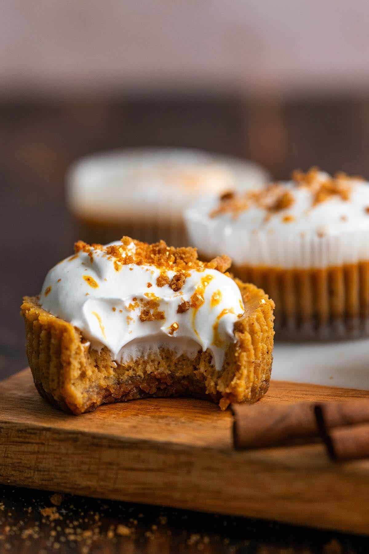 A close-up of a pumpkin pie cupcake on a wooden board, topped with whipped cream and crumbled nuts, with a bite taken out, conveying autumn warmth.