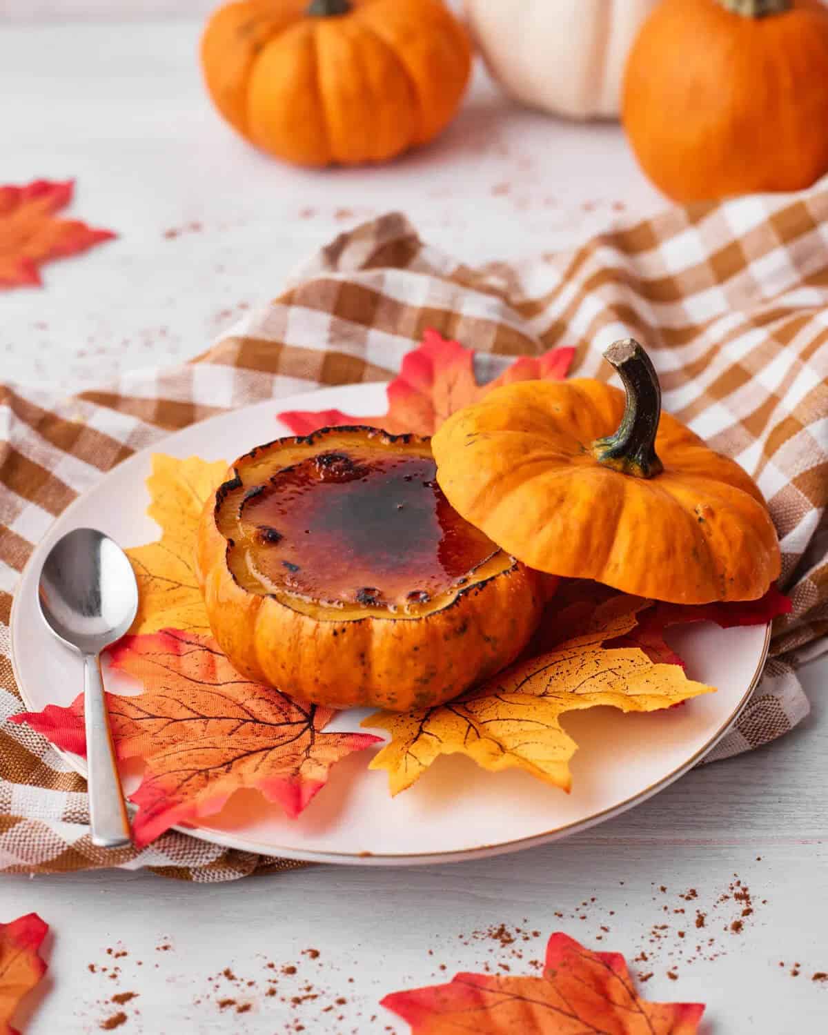 A pumpkin creme brûlée in a small pumpkin, placed on a white plate with autumn leaves and a spoon. Background has pumpkins and a checkered cloth. Cozy fall vibe.