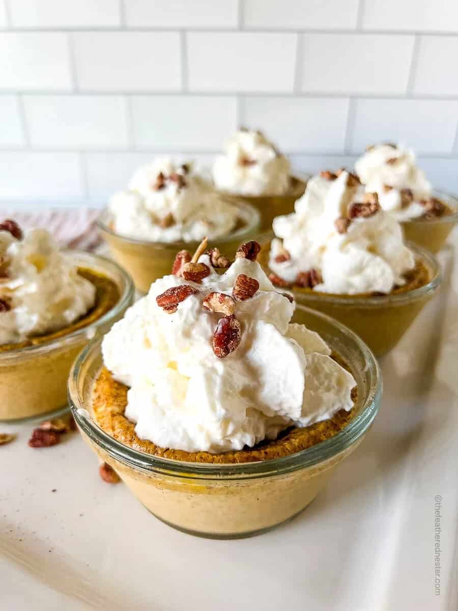 Mini pumpkin pies in glass dishes topped with whipped cream and pecans, set against a white tiled background. The scene feels cozy and inviting.