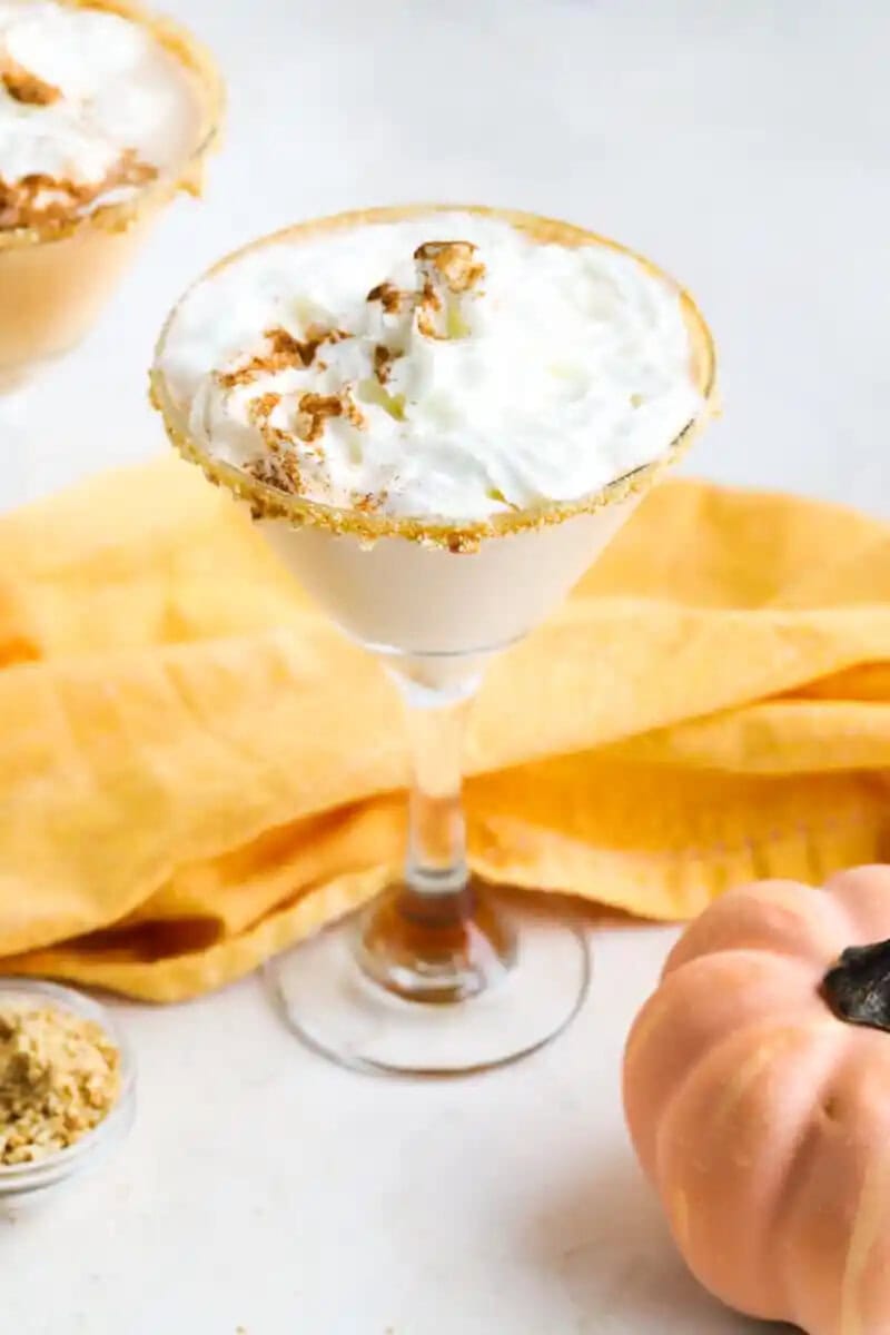 A creamy cocktail in a martini glass topped with whipped cream, cinnamon, and a graham cracker rim. Orange cloth and a small pumpkin create an autumn vibe.