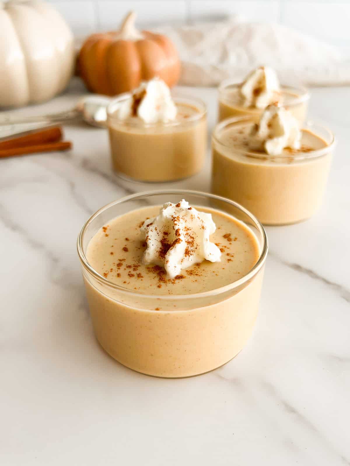 Four cups of creamy pumpkin pudding topped with whipped cream and a sprinkle of cinnamon are displayed on a marble surface, with pumpkins blurred in the background, evoking a cozy autumn vibe.