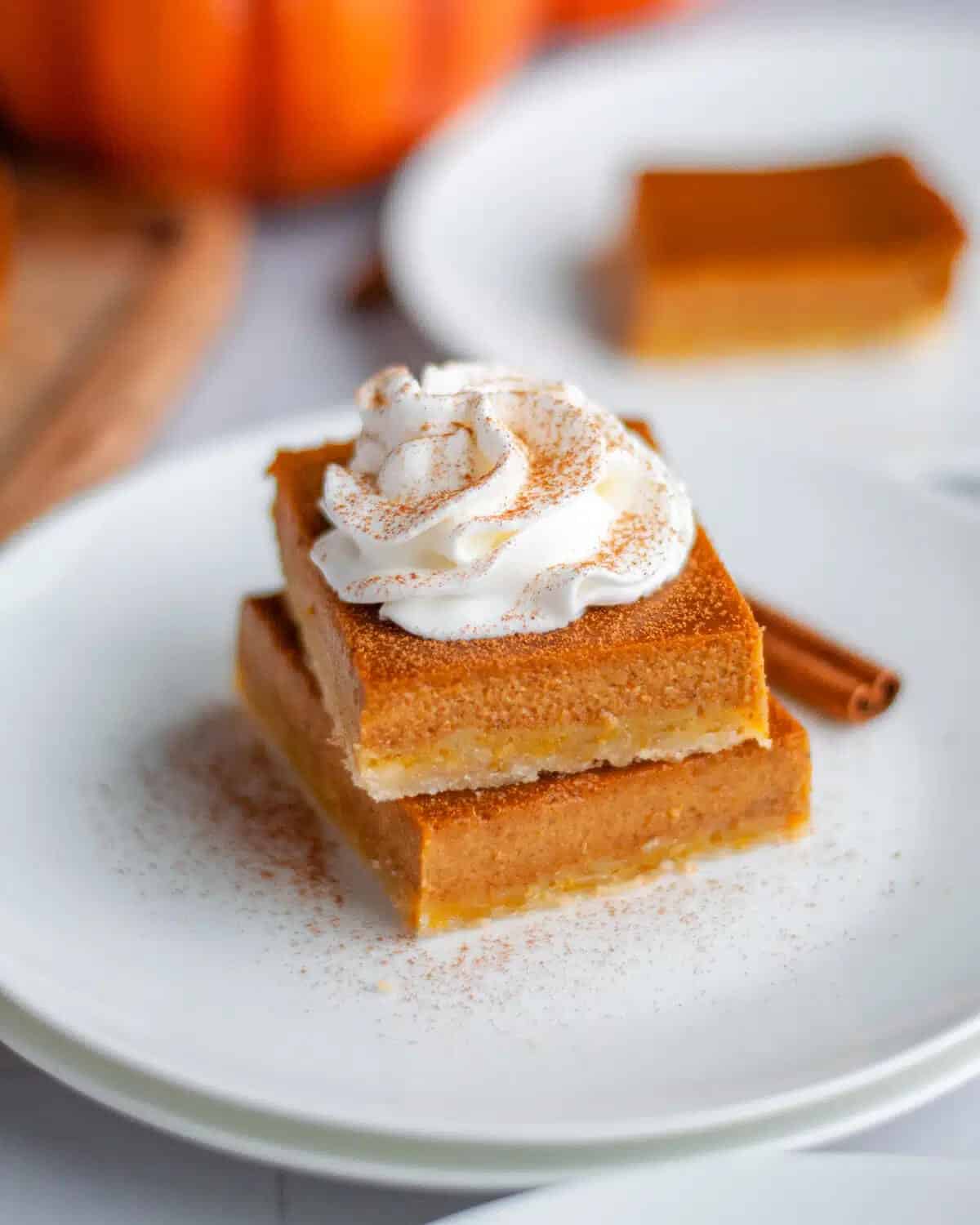 Two pumpkin pie bars stacked on a white plate, topped with whipped cream and cinnamon. A cinnamon stick is beside them, creating a cozy, autumn vibe.