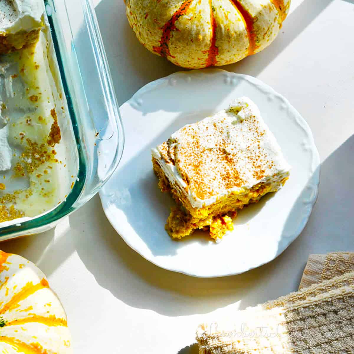 A plate with a slice of pumpkin dessert topped with cream, next to a glass baking dish with remaining portions. Decorative mini pumpkins nearby. Warm, inviting tone.