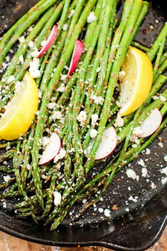 Grilled asparagus topped with crumbled feta, radish slices, and lemon wedges in a cast iron skillet, creating a fresh, vibrant dish.