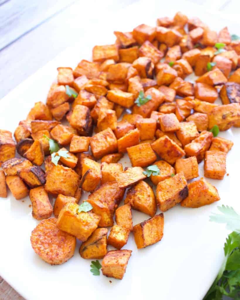 Crispy roasted sweet potato cubes scattered on a white platter, garnished with fresh cilantro. The bright orange hue suggests a savory, spiced flavor.
