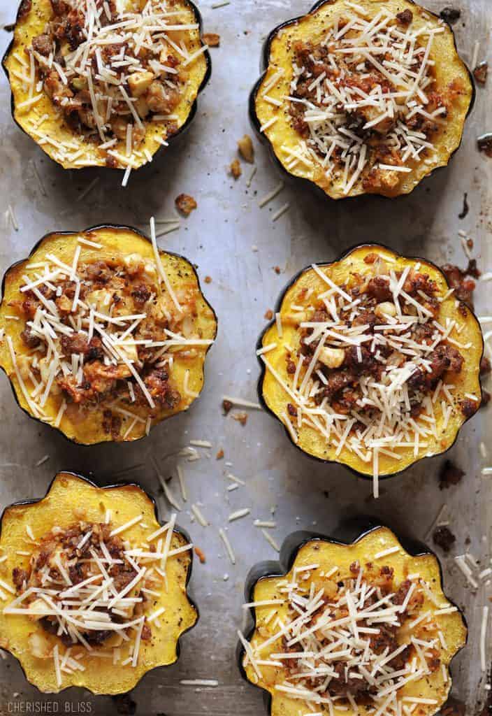 Six acorn squash halves filled with a savory stuffing topped with melted cheese are arranged on a baking sheet, suggesting a hearty and warm meal.