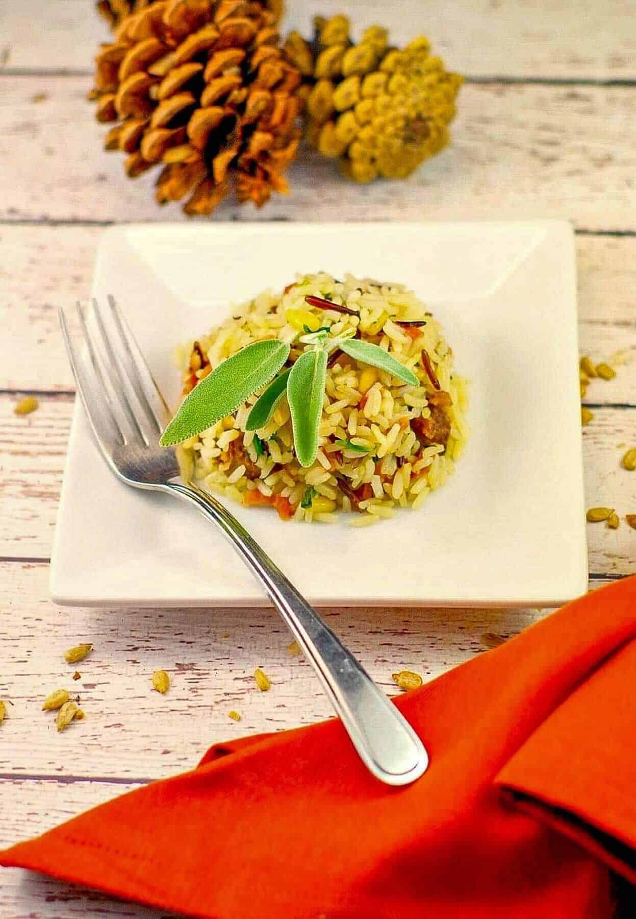A plate of rice garnished with sage on a square white dish, accompanied by a fork and orange napkin. Pine cones in the background add an autumnal feel.