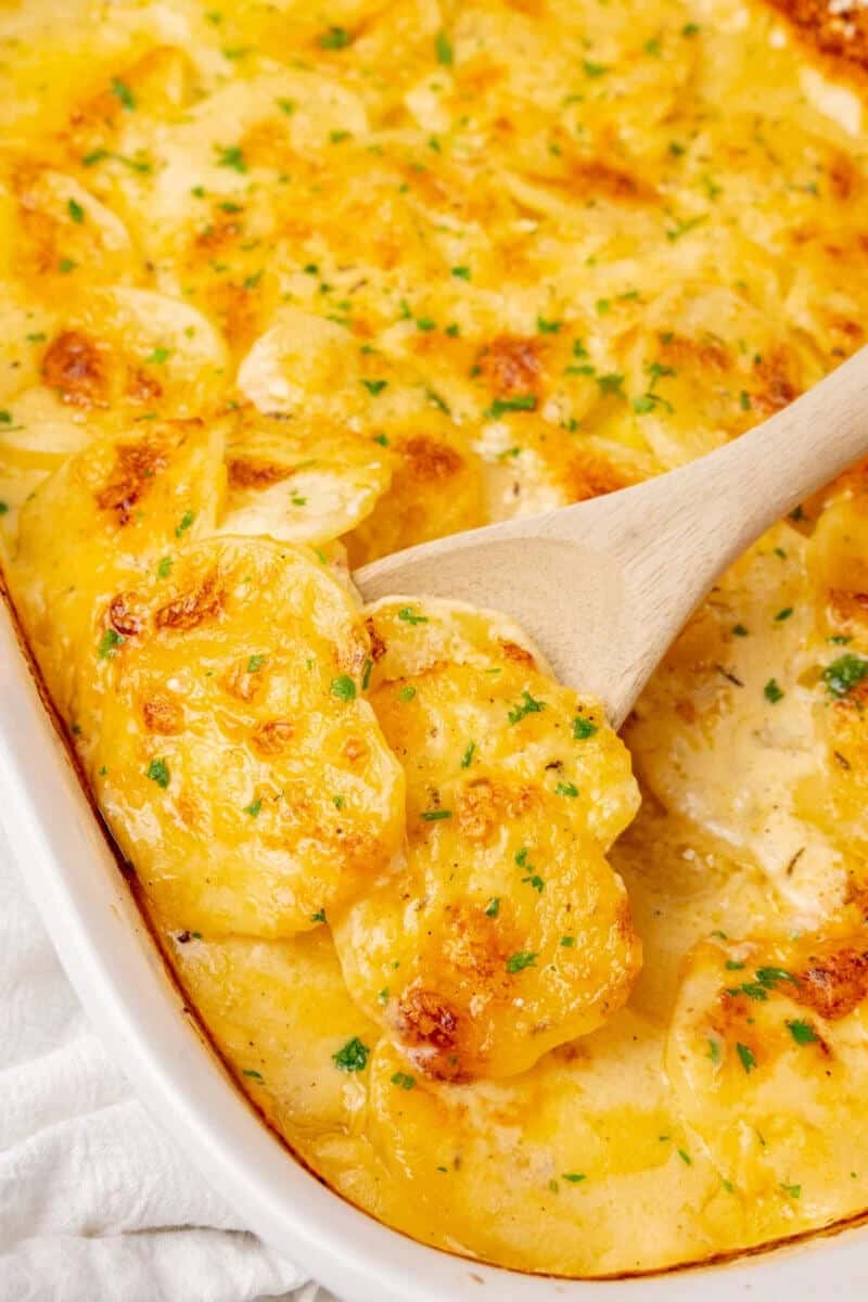Golden brown scalloped potatoes in a creamy sauce, garnished with parsley, being served from a baking dish with a wooden spoon. Warm, comforting feel.