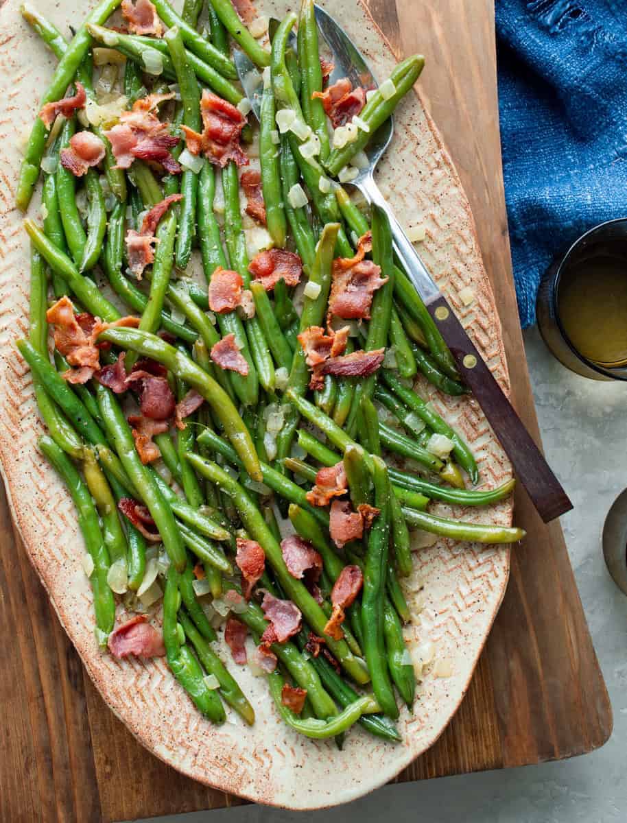 Oval platter with bright green beans mixed with crispy bacon pieces and chopped onions. Utensils and a blue napkin are nearby.